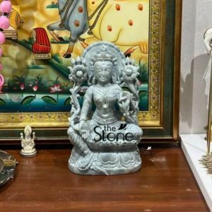 A small yet exquisite 7-inch Tara stone statue meticulously carved from greenstone is centrally displayed on a polished wooden surface likely a table or altar in a home setting. The statue depicts the Buddhist deity Tara revered as a Bodhisattva of compassion and action seated gracefully in a meditative posture possibly Vajrasana or a similar crossed-leg pose. The greenstone material gives the statue a beautiful muted greenish-gray hue with subtle natural variations emphasizing its authentic stone origin and delicate craftsmanship. Tara is richly adorned with intricate finely carved details representing traditional jewelry and spiritual attire. These include a magnificent multi-tiered crown ornate necklaces armlets, and bracelets all intricately rendered. Her slender torso is bare while her lower body is draped in robes with subtle folds. Her hands are in a mudra possibly the Varada Mudra or Abhaya Mudra holding what appear to be lotus stems or flowers. Behind her an elaborate carved halo or aura frames her figure featuring delicate floral patterns and rising lotus blossoms on either side symbolizing purity and enlightenment. Her serene face features gently closed or downcast eyes conveying deep meditation compassion and profound calm. The entire figure rests on a beautifully carved lotus pedestal. In the background a framed traditional Indian painting with vibrant yellows, reds and greens depicts a deity on a white bull, adding a rich cultural context. To the left of the statue a tiny white Ganesha figurine is visible. The warm wooden surface and surrounding elements create a sacred and aesthetically pleasing display making this greenstone Tara statue an ideal spiritual and decorative addition to a home in Mumbai embodying grace wisdom and compassion.