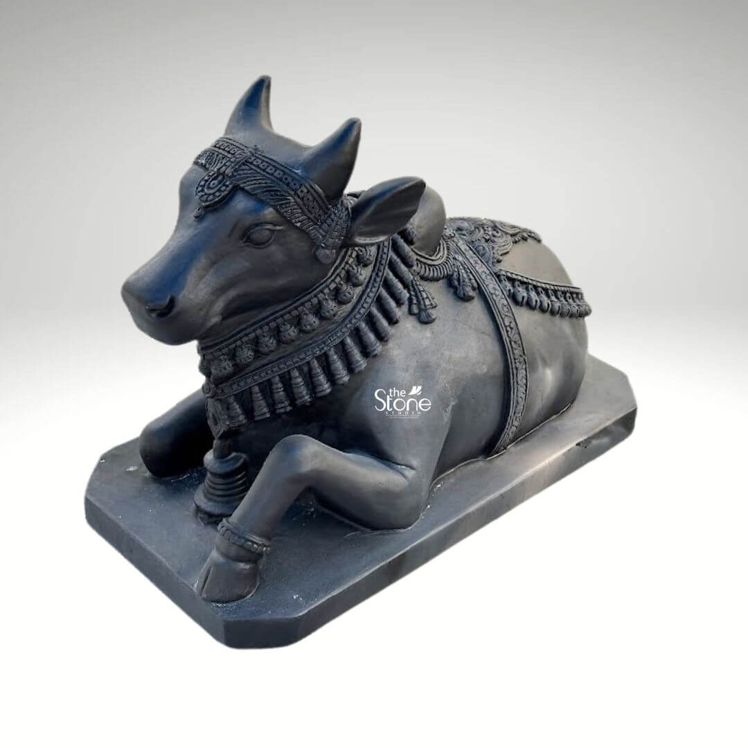 Black Nandi Bull Statue for Home 2.5ft 10 A majestic 2.5-foot traditional black stone sitting Nandi Bull statue is prominently featured against a clean neutral background emphasizing its intricate details and serene presence. This exquisite sculpture depicts Nandi the revered vahana of Lord Shiva in a classic reclining posture symbolizing unwavering patience devotion and strength making it ideal for an article discussing traditional sitting Nandi bull statues. The bull is shown lying down with its legs gracefully folded beneath its body its head turned slightly to the left looking forward with a calm and benevolent expression. The craftsmanship of the statue is exceptional showcasing the smooth deep black stone that beautifully accentuates Nandi's powerful yet serene form. The bull is richly adorned with meticulously carved traditional Indian jewelry and ceremonial embellishments. These include an elaborate crown or headpiece on its forehead intricately designed necklaces with dangling ornaments around its neck (including what appears to be a large bell or ghanta between its front legs and decorative blankets or saddle covers carved onto its back all integrated seamlessly into the stone. The details of the ornaments, such as beads bells and elaborate patterns are finely rendered enhancing the statue's divine appearance and cultural authenticity. A prominent hump is visible on its back adding to its realistic depiction. Nandi's facial features are finely sculpted with a tranquil and benevolent expression characterized by gentle eyes a strong snout and well-formed horns that rise gracefully from its head. The texture of the black stone, with its inherent depth and subtle sheen, gives the statue a timeless and dignified quality. The bull rests on a sturdy, rectangular black stone base which provides stability and a formal presentation. This 2.5-foot blackstone Nandi statue is not merely a decorative item but a powerful spiritual artifact inspiring deep devotion and bringing an auspicious presence to any home temple or sacred space.