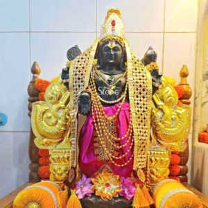 An eighteen-inch black stone Vishnu statue, adorned regally, is seated on an elaborate golden throne, suggesting its revered presence in a temple or shrine in Nashik, Maharashtra. The statue depicts Lord Vishnu with his traditional four arms, each holding a distinct attribute: the Sudarshana Chakra (discus) and Shankha (conch) are visible, subtly positioned behind his shoulders, while his primary hands are gracefully posed. He is draped in a vibrant, rich pink dhoti, adding a striking contrast to the dark stone and golden embellishments. A long, multi-layered golden bead necklace cascades down his chest, adding to his divine grandeur, complementing the intricate golden crown adorned with a red and white crest that sits majestically on his head. The Vishnu murti is further embellished with a rich, golden patterned shawl or stole that drapes over his shoulders and arms, adding texture and opulence to his attire. The black stone of the statue provides a beautiful canvas for the vibrant colors and gleaming gold, highlighting the deity's serene expression and traditional features. He is seated on a magnificent throne, characterized by its ornate golden carvings that feature stylized mythical creatures or motifs, possibly garudas or peacocks, on its armrests and back. The throne is made of a light-colored wood, possibly sandalwood, which is beautifully polished and enhances the golden elements. The base of the statue is surrounded by fresh pink and white flowers, scattered as offerings, signifying devotion and purity. Two plush, golden-yellow cushions with intricate designs and tassels flank the deity on the throne, suggesting comfort and reverence. The background features light-colored tiled walls, typical of a sacred inner sanctum, further emphasizing the statue's role as an object of worship. This image perfectly illustrates an article about a revered Vishnu stone statue in Nashik, Maharashtra, emphasizing its cultural and religious significance through its rich adornments, traditional setting, and the serene portrayal of the deity.