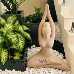 A serene three-foot off-white sandstone yoga statue is elegantly positioned in a modern indoor garden or relaxation space suggesting its presence in a stylish Dubai setting. The statue depicts a stylized figure in a classic yoga pose seated with legs crossed in a meditative posture and arms gracefully raised above the head with palms pressed together in an Anjali Mudra. The smooth light-colored sandstone gives the sculpture a contemporary and minimalist aesthetic highlighting its flowing lines and peaceful form. The statue's design is minimalist focusing on the silhouette and posture rather than intricate facial details creating a universal symbol of tranquility and meditation. The figure's form is simplified with gentle curves that convey a sense of calm and spiritual balance. The off-white hue of the sandstone contrasts beautifully with the surrounding elements making the statue a prominent focal point. The statue is placed on a bed of contrasting white and black pebbles creating a visually appealing foundation that accentuates its clean lines. Surrounding the statue are lush green plants in dark planters adding a touch of natural vibrancy to the scene. To the right, a modern staircase with white steps and sleek metal railings hints at a contemporary architectural environment. The overall composition evokes a sense of peace harmony, and modern elegance, making this yoga stone statue an ideal addition to a sophisticated interior design in Dubai perfectly suited for an article discussing such contemplative art pieces.