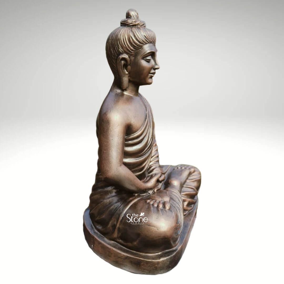 2 Feet Fibre Buddha Statue 2 A serene 2-foot fiber Buddha idol with a metallic bronze-like finish is gracefully presented in a side profile against a neutral background making it an ideal piece for home decor. The Buddha is depicted in the classic Dhyana Mudra seated in full lotus position with his hands resting gently in his lap palms upward symbolizing deep contemplation and enlightenment. His eyes are gently closed conveying profound inner peace and spiritual wisdom while a subtle smile graces his lips and his elongated earlobes are visible all characteristic features of traditional Buddha iconography. The lustrous finish of the fiber material made to resemble aged bronze adds a sense of timeless dignity and elegance to the sculpture. The detailing on the idol is meticulously rendered from the individual curls of his hair gathered in a prominent topknot to the graceful natural folds of his monastic robe that drape softly over his left shoulder and body. The side view emphasizes the harmonious proportions of the statue and the subtle contours of his form highlighting the artisan's skill. The bronze-like appearance effectively mimics the rich patina of an antique metal piece offering both aesthetic appeal and the practical benefits of durable fiber construction which is lighter yet robust. The Buddha idol rests on a simple low and subtly curved rectangular base which seamlessly integrates with the overall form providing stability without distracting from the main figure. This 2-foot fiber Buddha idol with its elegant bronze-like finish and peaceful demeanor embodies tranquility and spiritual grace making it a compelling subject for an article exploring aesthetically pleasing and durable Buddha statues suitable for enhancing the serene ambiance of any home.
