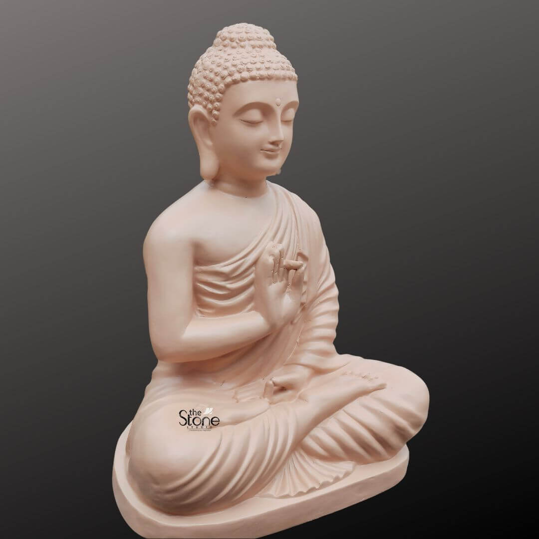 Buddha Statue for Zen Decor 2ft 2 A serene 2-foot fiber Buddha idol with a smooth light beige finish is elegantly presented in a slightly angled view against a dark gradient background making it an ideal piece for home decor. The Buddha is depicted in the Vitarka Mudra with his right hand raised palm outward and thumb and index finger touching to form a circle symbolizing the teaching and transmission of Buddhist doctrines. His left hand rests gently in his lap palms upward. Seated in full lotus position his eyes are gently closed conveying profound inner peace and spiritual wisdom while a subtle smile graces his lips and his elongated earlobes are visible all characteristic features of traditional Buddha iconography. The detailing on the idol is meticulously rendered from the individual curls of his hair gathered in a prominent topknot to the graceful natural folds of his monastic robe that drape softly over his left shoulder and body. The smooth matte finish of the fiber material gives the statue a natural stone-like appearanc perfectly aligning with various home aesthetics. The light beige color evokes a sense of calm and purity enhancing its contemplative presence. The fiber construction ensures that the statue is robust yet lighter than traditional stone offering both aesthetic appeal and practical versatility for different rooms in a home. The Buddha idol rests on a simple low and subtly curved rectangular base which seamlessly integrates with the overall form providing stability without distracting from the main figure. This 2-foot fiber Buddha idol with its elegant finish and peaceful demeanor embodies tranquility and spiritual grace serving as a captivating subject for an article exploring aesthetically pleasing and durable Buddha statues suitable for enhancing the serene and meditative ambiance of any home.
