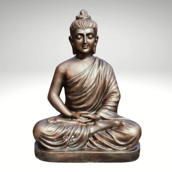 A serene 2-foot fiber Buddha statue finished with a metallic bronze-like coating is elegantly presented against a neutral background. The Buddha is depicted in the classic Dhyana Mudra seated in full lotus position with his hands resting gently in his lap palms upward symbolizing deep contemplation and enlightenment. His eyes are closed conveying profound inner peace while a gentle smile graces his lips and his elongated earlobes are visible all characteristic features of traditional Buddha iconography. The smooth lustrous finish of the fiber material made to resemble aged bronze adds a sense of timeless dignity to the sculpture. The detailing on the statue is meticulously rendered from the individual curls of his hair gathered in a topknot to the graceful folds of his monastic robe that drape naturally over his left shoulder and body. The light catches the contours of the statue highlighting the subtle musculature and the serene expression. The statue's appearance effectively mimics the rich patina of an antique bronze piece offering both aesthetic appeal and durability. The fiber construction allows for a lighter yet robust statue suitable for various settings. The Buddha statue rests on a simple low rectangular base which seamlessly integrates with the overall form providing stability without distracting from the main figure. This 2-foot fiber Buddha statue with its bronze-like finish, embodies tranquility and spiritual grace making it a compelling subject for an article exploring durable and aesthetically pleasing Buddha statues perfect for both indoor and outdoor contemplative spaces.