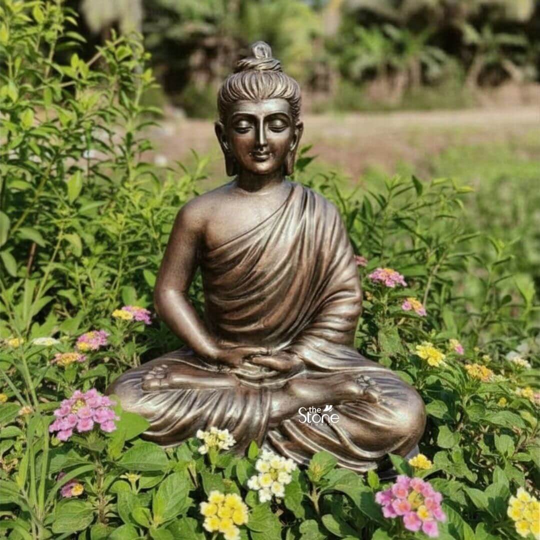 2 Feet Fibre Buddha Statue 3 A serene 2-foot fiber Buddha statue finished with a rich bronze-like patina is beautifully situated amidst a vibrant garden making it an inviting centerpiece for a foyer. The Buddha is depicted in the classic Dhyana Mudra seated in full lotus position with his hands gently resting in his lap palms upward symbolizing deep contemplation and enlightenment. His eyes are closed in profound peace a subtle smile graces his lips and his elongated earlobes are visible all characteristic features of traditional Buddha iconography. The lustrous aged bronze appearance of the fiber material lends the statue an aura of ancient wisdom and timeless serenity. The intricate details on the statue are meticulously rendered from the individual curls of his hair gathered in a topknot to the graceful natural folds of his monastic robe that drape softly over his left shoulder and body. The light plays across the contours of the statue highlighting its smooth surface and the dignified expression. The durable fiber construction ensures that the statue is robust yet lighter than traditional metal making it a versatile and appealing choice for a foyer where both aesthetic impact and practicality are important. The Buddha statue is surrounded by lush green foliage and a scattering of delicate pink and yellow flowers creating a natural and tranquil setting that enhances its spiritual presence. The soft focus of the background allows the statue to stand out as the primary focal point. This 2-foot fiber Buddha statue with its elegant bronze-like finish and peaceful demeano embodies tranquility and spiritual grace serving as a captivating subject for an article exploring aesthetically pleasing and durable Buddha statues suitable for welcoming guests in a foyer.