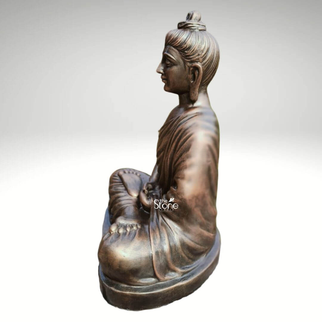 2 Feet Fibre Buddha Statue 4 A serene 2-foot fiber Buddha idol, with a metallic bronze-like finish is gracefully presented in a side profile against a neutral background making it an ideal piece for a shelf. The Buddha is depicted in the classic Dhyana Mudra seated in full lotus position with his hands resting gently in his lap palms upward symbolizing deep contemplation and enlightenment. His eyes are gently closed conveying profound inner peace and spiritual wisdom while a subtle smile graces his lips and his elongated earlobes are visible all characteristic features of traditional Buddha iconography. The lustrous finish of the fiber material made to resemble aged bronze adds a sense of timeless dignity and elegance to the sculpture. The detailing on the idol is meticulously rendered from the individual curls of his hair gathered in a prominent topknot to the graceful natural folds of his monastic robe that drape softly over his left shoulder and body. The side view emphasizes the harmonious proportions of the statue and the subtle contours of his form highlighting the artisan's skill. The bronze-like appearance effectively mimics the rich patina of an antique metal piece offering both aesthetic appeal and the practical benefits of durable fiber construction which is lighter yet robust making it suitable for display on a shelf. The Buddha idol rests on a simple, low and subtly curved rectangular base which seamlessly integrates with the overall form providing stability without distracting from the main figure. This 2-foot fiber Buddha idol with its elegant bronze-like finish and peaceful demeanor embodies tranquility and spiritual grace making it a compelling subject for an article exploring aesthetically pleasing and durable Buddha statues suitable for enhancing the serene ambiance of a shelf in any room.