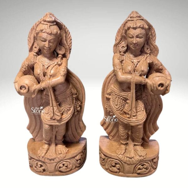 Two exquisite 2-foot sandstone Devdasi statues stand gracefully side-by-side ideal for garden decor. Each statue depicts a female figure a Devdasi adorned with traditional Indian attire and intricate jewelry including necklaces armlets, and bangles. Their serene faces are framed by beautifully sculpted hair and their slender forms convey elegance and movement. One hand of each statue delicately holds a spherical pot while the other hand gently rests on the pot or extends downwards suggesting an offering or a dance pose. The statues are carved from light-colored sandstone which brings out the delicate details of their garments and ornaments as well as the flowing lines of their drapery that extend behind them. Each figure stands on a circular intricately carved base featuring traditional patterns that enhance the overall aesthetic. The craftsmanship highlights the intricate details from the facial features to the intricate patterns on their attire showcasing the rich cultural heritage. The identical nature of the two statues positioned as a pair emphasizes their symmetry and allows for a balanced aesthetic when used in garden settings. This pair of 2ft Sandstone Devdasi statues truly embodies classical Indian artistry and cultural heritage making them perfect artistic additions for enhancing garden aesthetics and creating a focal point in any outdoor space.
