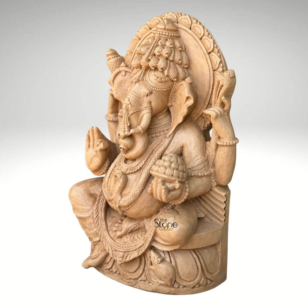 3ft Sitting Ganesha Statue 2 A magnificent 3-foot sandstone Ganesha statue carved from rich brown sandstone is prominently displayed in a three quarter side view against a plain background making it a captivating subject for an article about such sculptures. Lord Ganesha is depicted in a traditional seated posture with his iconic elephant head adorned by an elaborate crown and intricate carvings detailing his large ears and trunk. He is shown with four arms holding a conch shell and an axe in his upper hands, a modak (sweet) in one lower hand, and his other lower hand raised in a gesture of blessing symbolizing protection and grace. The statue is richly adorned with necklaces armlets and a sacred thread draped across his plump belly all meticulously carved from the sandstone. The large ears and expressive eyes give the Ganesha statue a benevolent and wise demeanor. Behind his head a circular halo-like structure is intricately carved with traditional patterns further accentuating his divine aura. The craftsmanship is evident in every detail from the delicate folds of his dhoti to the robust and powerful rendering of his form all enhanced by the warm natural hue and texture of the brown sandstone. Ganesha is seated on a lotus pedestal which is also intricately carved with floral motifs and drapery providing a stable and aesthetically pleasing foundation. The brown sandstone's natural characteristics highlight the deep textures and intricate patterns of the carving making every detail stand out with an ancient and earthy feel. This 3-foot sandstone Ganesha statue truly embodies spiritual reverence and artistic excellence serving as a powerful and beautiful representation of the beloved deity.