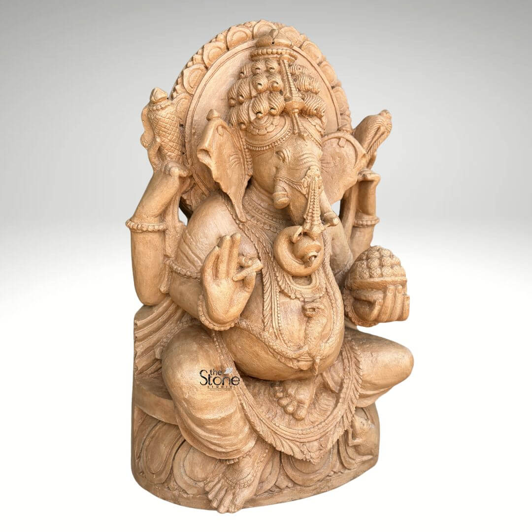 3ft Sitting Ganesha Statue 4 A magnificent 3-foot brown sandstone Ganesha statue is prominently displayed in a slightly angled front-facing view against a plain background making it a captivating subject for an article about such sculptures. Lord Ganesha is depicted in a traditional seated posture with his iconic elephant head adorned by an elaborate crown and intricate carvings detailing his large ears and trunk. He is shown with four arms holding a conch shell and an axe in his upper hands a modak in one lower hand and his other lower hand raised in a gesture of blessing symbolizing protection and grace. The statue is richly adorned with necklaces armlets and a sacred thread draped across his plump belly all meticulously carved from the brown sandstone. The large ears and expressive eyes give the Ganesha statue a benevolent and wise demeanor. Behind his head a circular halo-like structure is intricately carved with traditional patterns further accentuating his divine aura. The craftsmanship is evident in every detais from the delicate folds of his dhoti to the robust and powerful rendering of his form all enhanced by the warm natural hue and textured finish of the brown sandstone. Ganesha is seated on a lotus pedestal which is also intricately carved with floral motifs and drapery providing a stable and aesthetically pleasing foundation. The brown sandstone's natural characteristics highlight the deep textures and intricate patterns of the carving, making every detail stand out with an ancient and earthy feel. This 3-foot brown sandstone Ganesha statue truly embodies spiritual reverence and artistic excellence serving as a powerful and beautiful representation of the beloved deity.
