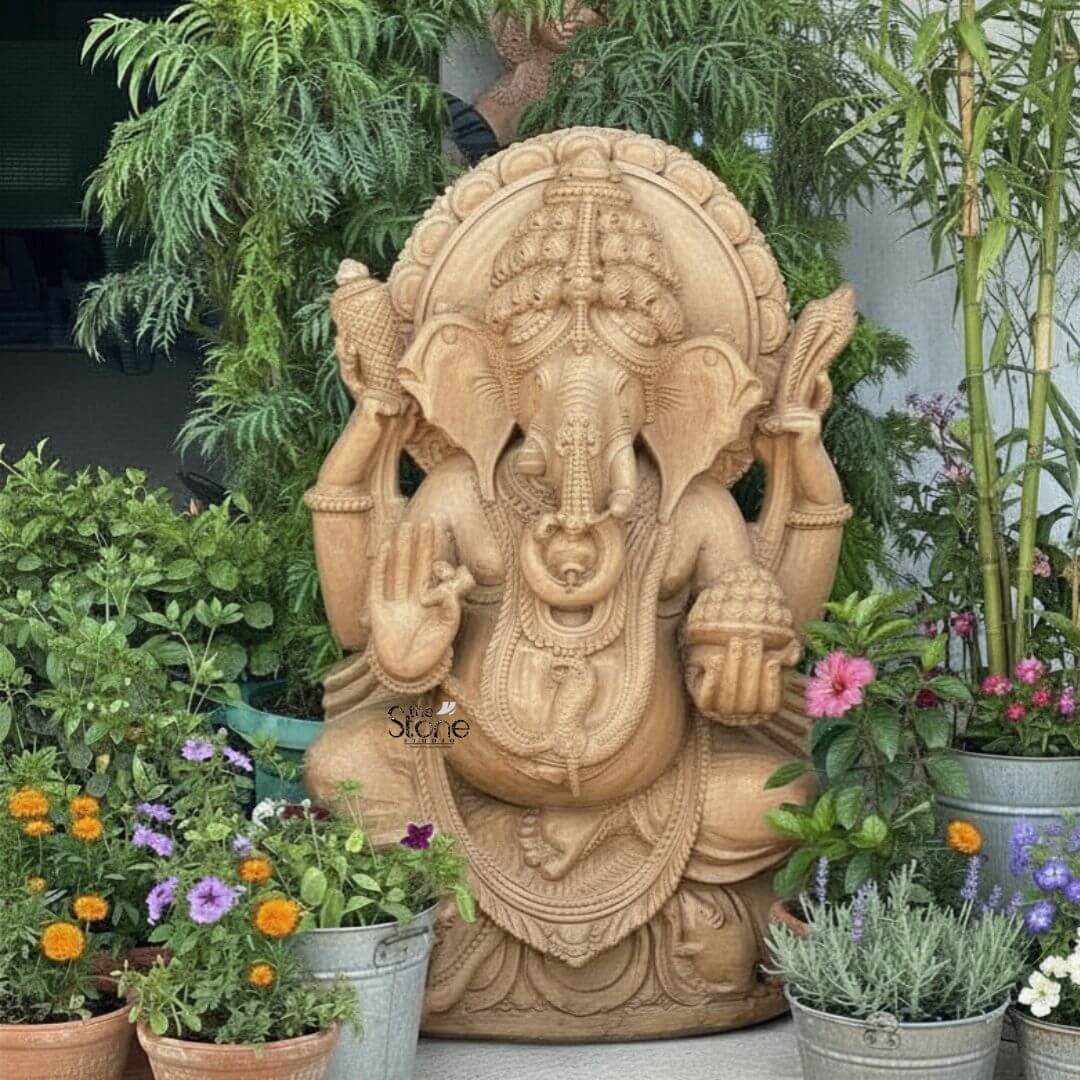 3ft Sitting Ganesha Statue 3 A magnificent 3-foot Lord Ganesha sandstone statue carved from rich brown sandstone is prominently displayed in a lush outdoor setting making it a captivating subject for an article about such sculptures. Lord Ganesha is depicted in a traditional seated posture with his iconic elephant head adorned by an elaborate crown and intricate carvings detailing his large ears and trunk. He is shown with four arms, holding a conch shell and an axe in his upper hands a modak in one lower hand, and his other lower hand raised in a gesture of blessing symbolizing protection and grace. The statue is richly adorned with necklaces armlets and a sacred thread draped across his plump belly all meticulously carved from the sandstone. The large ears and expressive eyes give the Ganesha statue a benevolent and wise demeanor. Behind his head a circular halo like structure is intricately carved with traditional patterns further accentuating his divine aura. The craftsmanship is evident in every detail from the delicate folds of his dhoti to the robust and powerful rendering of his form all enhanced by the warm natural hue and textured finish of the brown sandstone, which blends harmoniously with the garden. Ganesha is seated on a lotus pedestal which is also intricately carved with floral motifs and drapery providing a stable and aesthetically pleasing foundation. The statue is surrounded by vibrant potted plants and greenery including tall bamboo shoots various flowers in shades of pink orange, and purple and lush foliage all adding to the serene and inviting atmosphere. The natural light highlights the details of the carving and the textures of the plants creating a harmonious and spiritual scene making this 3-foot Lord Ganesha sandstone statue a powerful and beautiful representation of the beloved deity.