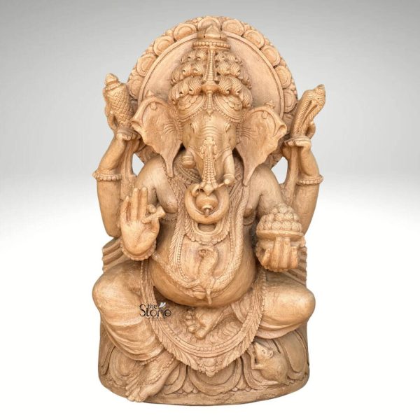 A magnificent 3-foot sitting Ganesha statue carved from rich brown sandstone is prominently displayed against a plain background making it a captivating subject for an article about such sculptures. Lord Ganesha is depicted in a traditional seated posture with his iconic elephant head adorned by an elaborate crown and intricate carvings detailing his large ears and trunk. He is shown with four arms holding a conch shell and an axe in his upper hands a modak in one lower hand and his other lower hand raised in a gesture of blessing symbolizing protection and grace. The statue is richly adorned with necklaces armlets and a sacred thread draped across his plump belly all meticulously carved from the sandstone. The large ears and expressive eyes give the Ganesha statue a benevolent and wise demeanor. Behind his head a circular halo-like structure is intricately carved with traditional patterns further accentuating his divine aura. The craftsmanship is evident in every detail from the delicate folds of his dhoti to the robust and powerful rendering of his form, all enhanced by the warm natural hue of the brown sandstone. Ganesha is seated on a lotus pedestal which is also intricately carved with floral motifs and drapery providing a stable and aesthetically pleasing foundation. The brown sandstone's natural texture and color highlight the deep textures and intricate patterns of the carving making every detail stand out with an ancient and earthy feel. This 3-foot sitting Ganesha statue truly embodies spiritual reverence and artistic excellence serving as a powerful and beautiful representation of the beloved deity.