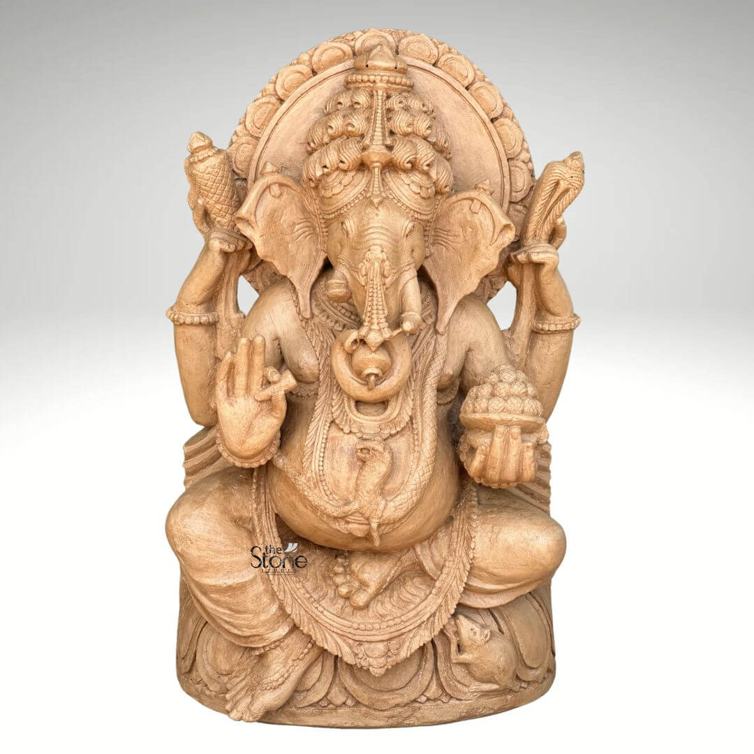 3ft Sitting Ganesha Statue 1 A magnificent 3-foot sitting Ganesha statue carved from rich brown sandstone is prominently displayed against a plain background making it a captivating subject for an article about such sculptures. Lord Ganesha is depicted in a traditional seated posture with his iconic elephant head adorned by an elaborate crown and intricate carvings detailing his large ears and trunk. He is shown with four arms holding a conch shell and an axe in his upper hands a modak in one lower hand and his other lower hand raised in a gesture of blessing symbolizing protection and grace. The statue is richly adorned with necklaces armlets and a sacred thread draped across his plump belly all meticulously carved from the sandstone. The large ears and expressive eyes give the Ganesha statue a benevolent and wise demeanor. Behind his head a circular halo-like structure is intricately carved with traditional patterns further accentuating his divine aura. The craftsmanship is evident in every detail from the delicate folds of his dhoti to the robust and powerful rendering of his form, all enhanced by the warm natural hue of the brown sandstone. Ganesha is seated on a lotus pedestal which is also intricately carved with floral motifs and drapery providing a stable and aesthetically pleasing foundation. The brown sandstone's natural texture and color highlight the deep textures and intricate patterns of the carving making every detail stand out with an ancient and earthy feel. This 3-foot sitting Ganesha statue truly embodies spiritual reverence and artistic excellence serving as a powerful and beautiful representation of the beloved deity.