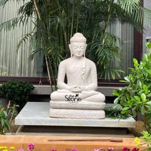 A serene 3-foot Stone Buddha Statue meticulously carved from white sandstone is gracefully positioned outdoors possibly in a garden or courtyard in Bangalore radiating peace and tranquility. The Buddha is depicted in a traditional meditative posture with both hands resting in his lap palms facing upwards signifying profound contemplation and inner calm. His face carries a tranquil expression with gently closed eyes and a subtle smile embodying deep spiritual focus and enlightenment. The ushnisha and tightly curled hair are characteristic features along with elongated earlobes. The white sandstone material gives the statue a pristine and luminous quality highlighting its smooth contours and intricate details. The Buddha is draped in a simple flowing monastic robe beautifully sculpted with delicate pleats that suggest the softness of fabric. He sits on a multi-tiered lotus base also carved from white sandstone which provides a symbolic and aesthetically pleasing foundation. This base rests upon a larger rectangular light-colored stone slab further elevating the statue and creating a designated sacred space. The light color of the sandstone contrasts harmoniously with the vibrant green foliage surrounding it enhancing its serene presence. The statue is set against a lush backdrop of various green plants including tall bamboo or similar slender-leafed plants directly behind it and dense bushes with broad leaves on either side. A window with light-colored curtains is visible in the background suggesting a nearby building blending the natural elements with a touch of architectural presence. In the foreground a bed of colorful flowers including pink and yellow blooms adds a vibrant touch of nature. This image perfectly illustrates an article about 3ft Stone Buddha Statues particularly white sandstone pieces in locations like Bangalore showcasing how such a statue can be a central element in creating a tranquil outdoor sanctuary embodying peace, enlightenment and harmonious integration with nature.
