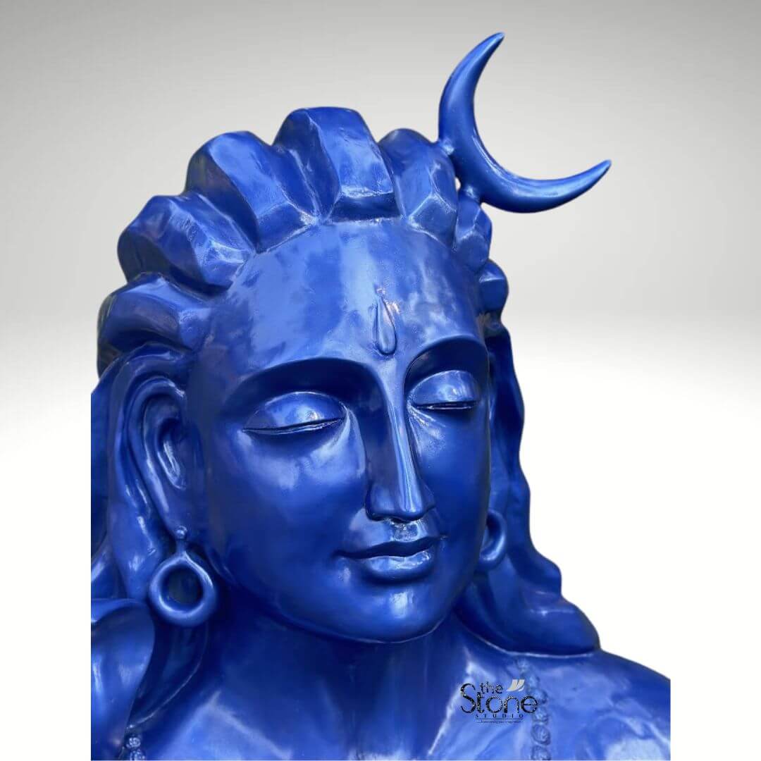 Blue Adiyogi Shiva Statue 4ft 11 A striking close-up of a 4-foot fiber Adiyogi Shiva bust statue is prominently displayed focusing on the serene face and matted locks making it an evocative subject for an article about such sculptures. Lord Shiva is depicted with closed eyes in deep meditation conveying profound inner peace and wisdom. His iconic third eye is subtly marked on his forehead and a prominent crescent moon gracefully adorns his matted locks which are styled upwards and flow around his head symbolizing his ascetic nature. The entire statue is rendered in a vibrant deep blue color characteristic of Shiva's divine complexion. Shiva's ears are adorned with traditional large ring earrings visible on the side of his head. The intricate detailing on his hair and facial features showcases remarkable artistry. The smooth glossy finish of the fiber material enhances the statue's vibrant blue hue making it a visually captivating piece that evokes both ancient reverence and modern artistic interpretation. The close-up view highlights the delicate contours of his face the subtle smile and the peaceful expression drawing the viewer into a state of contemplation. The fiber construction ensures the statue is durable yet relatively lightweight suitable for various indoor and outdoor settings. This 4-foot fiber Adiyogi Shiva bust statue truly embodies spiritual power and artistic grace with its intense blue color adding to its mystique. Its commanding yet tranquil presence makes it a powerful symbol of yoga and ultimate liberation perfect for inspiring meditation and reverence.