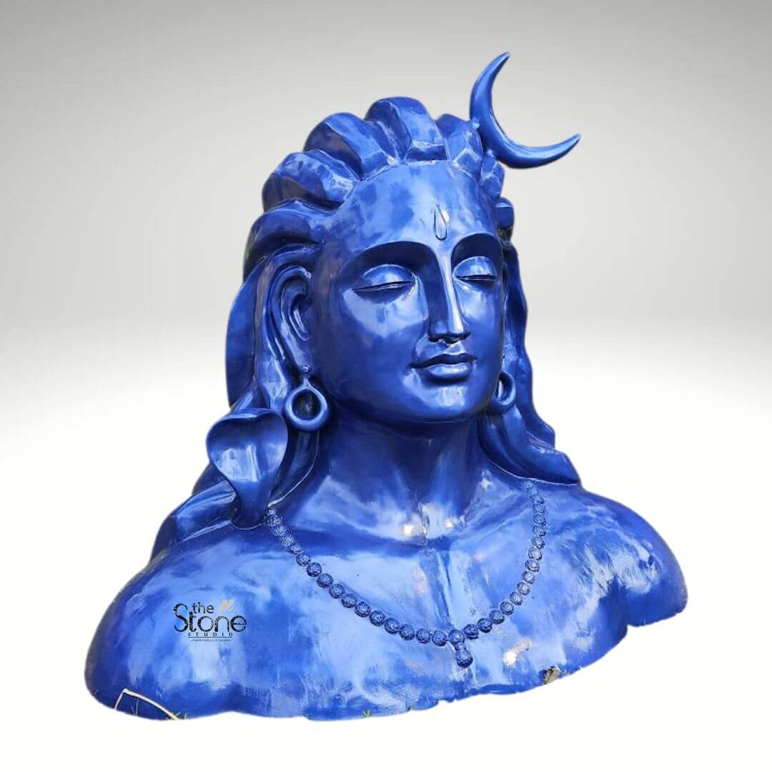 Blue Adiyogi Shiva Statue 4ft 2 A striking 4-foot fiber statue of the Adiyogi Shiva bust is prominently displayed against a plain background making it an evocative piece for home decor. The sculpture captures the upper torso and head of Lord Shiva in his Adiyogi form depicted with closed eyes in deep meditation conveying profound inner peace and wisdom. His iconic third eye is subtly marked on his forehead and a prominent crescent moon gracefully adorns his matted locks which are styled upwards and flow around his shoulders symbolizing his ascetic nature. The entire statue is rendered in a vibrant deep blue color characteristic of Shiva's divine complexion. Shiva's ears are adorned with traditional large ring earrings and a detailed serpent is subtly carved around his neck along with a beaded necklace adding to his divine iconography. The intricate detailing on his hair ornaments and facial features showcases remarkable artistry. The smooth glossy finish of the fiber material enhances the statue's vibrant blue hue making it a visually captivating piece that evokes both ancient reverence and modern artistic interpretation. The robust fiber construction ensures the statue is durable yet relatively lightweight suitable for various indoor and outdoor settings within a home. The statue's bust-like form with its broad shoulders and serene countenance exudes a powerful yet tranquil presence making it a compelling focal point in any room or garden. This 4-foot fiber Adiyogi Shiva statue truly embodies spiritual power and artistic grace, with its intense blue color adding to its mystique. Its commanding yet peaceful presence makes it a powerful symbol of yoga and ultimate liberation perfect for inspiring contemplation and reverence in a home environment.