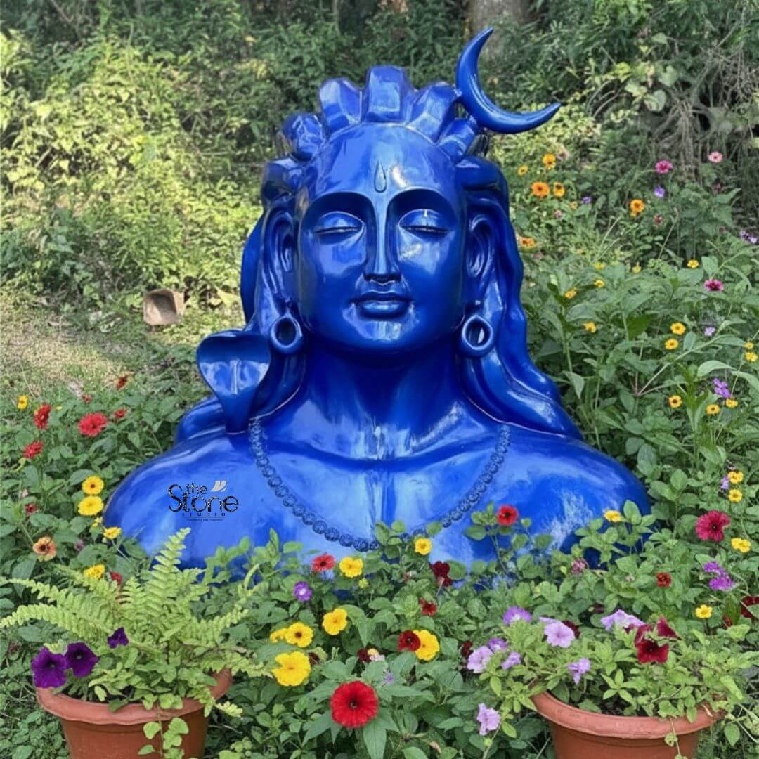 Blue Adiyogi Shiva Statue 4ft 3 A striking 4-foot fiber statue of the Blue Adiyogi Shiva bust is beautifully nestled in a vibrant outdoor garden setting making it an evocative piece for outdoor decor. The sculpture captures the upper torso and head of Lord Shiva in his Adiyogi form depicted with closed eyes in deep meditation conveying profound inner peace and wisdom amidst nature. His iconic third eye is subtly marked on his forehead and a prominent crescent moon gracefully adorns his matted locks which are styled upwards and flow around his shoulders symbolizing his ascetic nature. The entire statue is rendered in a vibrant, deep blue color characteristic of Shiva's divine complexion. Shiva's ears are adorned with traditional large ring earrings, and a detailed serpent is carved around his neck, along with a beaded necklace, adding to his divine iconography. The intricate detailing on his hair, ornaments and facial features showcases remarkable artistry. The smooth glossy finish of the fiber material enhances the statue's vibrant blue hue making it a visually captivating piece that contrasts beautifully with the surrounding greenery and flowers. The robust fiber construction ensures the statue is durable and weather-resistant perfectly suited for outdoor display. The statue is surrounded by lush green foliage and a profusion of colorful flowers including reds yellows, and purples, some in terracotta pots in the foreground, creating a harmonious and spiritual garden tableau. The natural light illuminates the statue emphasizing its serene expression and the vibrant blue. This 4-foot fiber Adiyogi Shiva statue truly embodies spiritual power and artistic grace with its intense blue color adding to its mystique making it a powerful focal point for an article exploring such statues for outdoor decor.