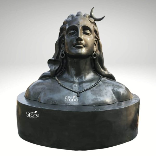 A majestic fiber Adiyogi Shiva Statue with an integrated pedestal is presented against a plain light grey background emphasizing its powerful and serene presence. This impressive sculpture depicts the bust of Lord Shiva the Adiyogi with a profoundly meditative expression. His eyes are gently closed conveying deep introspection and spiritual calm. Characteristic features include the crescent moon adorning his matted locks, a prominent third eye on his forehead and large earlobes adorned with simple ring earrings. A coiled serpent is sculpted around his neck symbolizing his mastery over death and time. Shiva's matted hair flows gracefully adding to his ascetic yet divine appearance. He wears a simple yet elegant necklace of rudraksha beads which drapes across his powerful chest. The entire sculpture is finished in a uniform dark almost metallic grey or black color giving it a monolithic and ancient stone-like quality despite being made of fiber. This finish highlights the smooth contours and intricate details of the carving from the subtle smile on his lips to the texture of his hair and the serpent. The use of fiber allows for intricate details and a polished finish while being lighter than traditional stone. The bust of Shiva seamlessly merges with a robust cylindrical pedestal forming a single integrated piece. The pedestal has a smooth dark surface providing a sturdy and dignified base that elevates the deity. This design choice contributes to the statue's commanding presence making it an ideal focal point for a home meditation center or outdoor space. This image effectively illustrates an article about Adiyogi Shiva Statues with pedestals particularly those crafted from fiber emphasizing its artistic merit spiritual significance and its ability to inspire peace and devotion.
