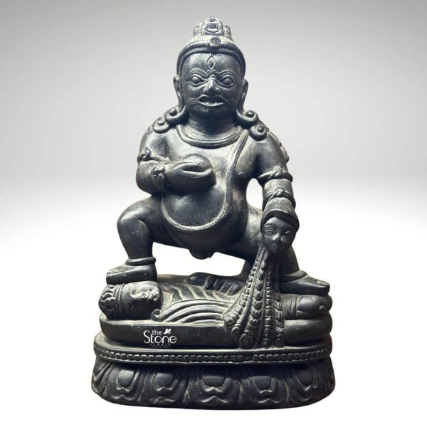 A commanding 9-inch Jambhala statue meticulously carved from black stone is prominently displayed against a neutral background highlighting its intricate details and powerful presence. The deity is depicted in a squatting or crouching posture exuding a sense of robust strength and determination. Jambhala's facial features are distinct with a stern expression wide eyes and a slight smirk conveying both intensity and benevolence. The dark, polished black stone accentuates the statue's muscular form, the folds of his belly and the rich textures of his attire and ornamentation. Jambhala is shown holding a small round object in his left hand traditionally a jewel or fruit symbolizing wealth and prosperity. His right hand rests on a mongoose that is spewing jewels a classic attribute representing his ability to bestow riches and eliminate poverty. The detailed carving extends to his headdress armlets and necklace all intricately rendered in the black stone. The statue rests on a beautifully sculpted lotus pedestal a common motif in Buddhist and Hindu iconography symbolizing purity and spiritual awakening despite being associated with material wealth. This image is ideally suited for an article discussing black stone Jambhala statues showcasing a prime example for collectors or those seeking symbols of prosperity for their homes or altars. The 9-inch size makes it a significant yet manageable piece and the black stone material lends it a timeless imposing quality. The detailed depiction of Jambhala's attributes and powerful posture directly conveys its connection to wealth protection, and spiritual abundance making it a compelling subject for the article's focus.