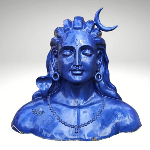 A striking 4 foot fiber statue of the Blue Adiyogi Shiva is prominently displayed against a plain background exuding a powerful and serene presence. The sculpture captures the upper torso and head of Lord Shiva in his Adiyogi form depicted with closed eyes in deep meditation conveying profound inner peace and wisdom. His iconic third eye is subtly marked on his forehead and a crescent moon gracefully adorns his matted locks which are styled upwards and flow around his shoulders symbolizing his ascetic nature. The entire statue is rendered in a vibrant deep blue color characteristic of Shiva's divine complexion. Shiva's ears are adorned with traditional large ring earrings and a detailed serpent is carved around his neck along with a beaded necklace adding to his divine iconography. The intricate detailing on his hair ornaments and facial features showcases remarkable artistry. The smooth glossy finish of the fiber material enhances the statue's vibrant blue hue making it a visually captivating piece that evokes both ancient reverence and modern artistic interpretation. The fiber construction ensures the statue is durable yet relatively lightweight suitable for various settings. The statue's bust-like form rises from what appears to be a natural grassy base, suggesting an outdoor installation. This 4-foot fiber Blue Adiyogi Shiva statue truly embodies spiritual power and artistic grace making it a compelling subject for an article exploring such statues and their profound significance as a symbol of yoga and ultimate liberation.