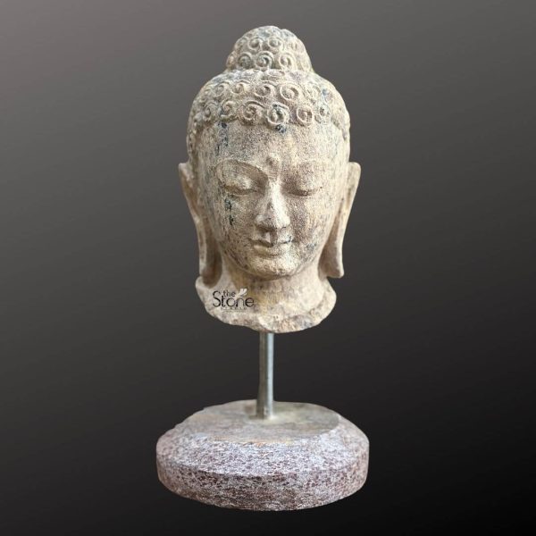 A serene 12-inch Buddha head statue meticulously carved from brown sandstone is elegantly displayed on a metal stand against a dark neutral background. The Buddha's head features finely sculpted details including closed or downcast eyes that convey deep inner peace and meditation a gentle smile and elongated earlobes indicative of his renunciate life. The brown sandstone exhibits a subtle textured finish with natural variations specks of darker inclusions and hints of weathering adding to its authentic ancient and earthy appeal. The hair is intricately depicted as tight small curls culminating in a prominent ushnisha at the top symbolizing his spiritual wisdom and enlightenment. The graceful contours of the face the delicate lines of the eyelids and the overall calm expression radiate a sense of profound tranquility and introspection. The 12-inch head is securely mounted on a slender metal rod that rises from a sturdy round stone base. This base has a more pronounced rough texture and a slightly richer reddish-brown hue providing a stable and contrasting foundation for the delicate head sculpture. This image is ideally suited for an article discussing Buddha head statues on stands showcasing a classic and aesthetically pleasing 12-inch brown sandstone example perfect for home decor meditation spaces, or as a focal point in a serene environment. The combination of the tranquil Buddha head the natural stone material with its warm tones and the elegant stand makes it a versatile piece that merges spiritual significance with artistic beauty. It effectively conveys a sense of calm and enlightenment inviting mindfulness and contemplation into any space it adorns.