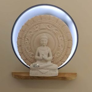 A captivating Buddha sculpture, crafted from off-white sandstone is elegantly displayed on a minimalist wooden shelf against a neutral-toned wall. The statue features the Buddha in a serene Dhyana Mudra with hands gently resting in his lap emanating profound peace and spiritual calm. The pristine off-white sandstone beautifully highlights the smooth contours of his form and the intricate details of his serene expression creating a focal point of tranquility. The base of the statue is adorned with subtle lotus petal carvings adding to its sacred symbolism. Behind the Buddha a larger circular halo or back panel also made of off-white sandstone provides a magnificent backdrop. This halo is richly embellished with multiple concentric rings of meticulously carved traditional motifs including stylized lotus patterns and delicate floral designs. What makes this display particularly striking is the modern circular frame possibly metal that encircles the sandstone halo integrated with a soft glowing LED light strip. This backlighting creates a captivating aura around the Buddha enhancing its divine presence and adding a contemporary touch to the traditional sculpture. This image is perfectly suited for an article discussing Buddha sculptures particularly in regions like Surat, Gujarat highlighting their aesthetic appeal and spiritual significance as home decor. The off-white sandstone statue, with its combined traditional craftsmanship and modern backlit display showcases a versatile piece that can elevate any interior space. It represents a harmonious blend of ancient artistry and contemporary design making it a powerful symbol of peace and a sophisticated decorative element for modern homes.