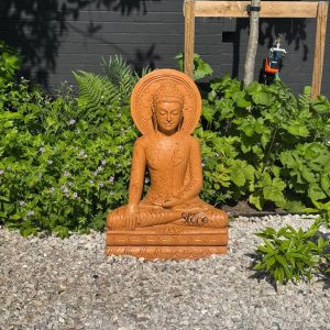 A serene 3-foot Buddha statue crafted from warm-toned sandstone sits gracefully in a contemplative pose within a verdant garden setting likely in Walton on Thames London as suggested by its article context. The Buddha is depicted with closed eyes a gentle smile and elongated earlobes conveying a sense of profound peace and inner tranquility. His hands rest in his lap in the Dhyana Mudra a gesture of meditation and focus which is characteristic of statues designed for calming environments. The natural texture of the sandstone is visible offering an earthy and grounded feel to the sculpture perfect for an outdoor spiritual space. The statue is positioned on a rectangular base also made of sandstone which rests on a bed of light-colored gravel. Surrounding the Buddha is a lush array of green plants including ferns flowering ground cover with small purple blossoms and larger leafy bushes creating a natural and harmonious backdrop. In the background a dark fence and the base of a tree are visible along with what appears to be a small outdoor spigot or watering device indicating a residential or cultivated garden environment typical of areas like Walton on Thames. The interplay of light and shadow subtly highlights the contours of the Buddha's face and robes, enhancing its spiritual presence. This 3-foot sandstone Buddha statue serves as a focal point radiating calm amidst the natural growth of the garden. Its size and material make it a suitable piece for a personal meditation space or a decorative element in a garden in London aligning perfectly with an article discussing Buddha Statue in Walton on Thames emphasizing both its artistic craftsmanship and its potential role in fostering tranquility within an urban or suburban landscape.