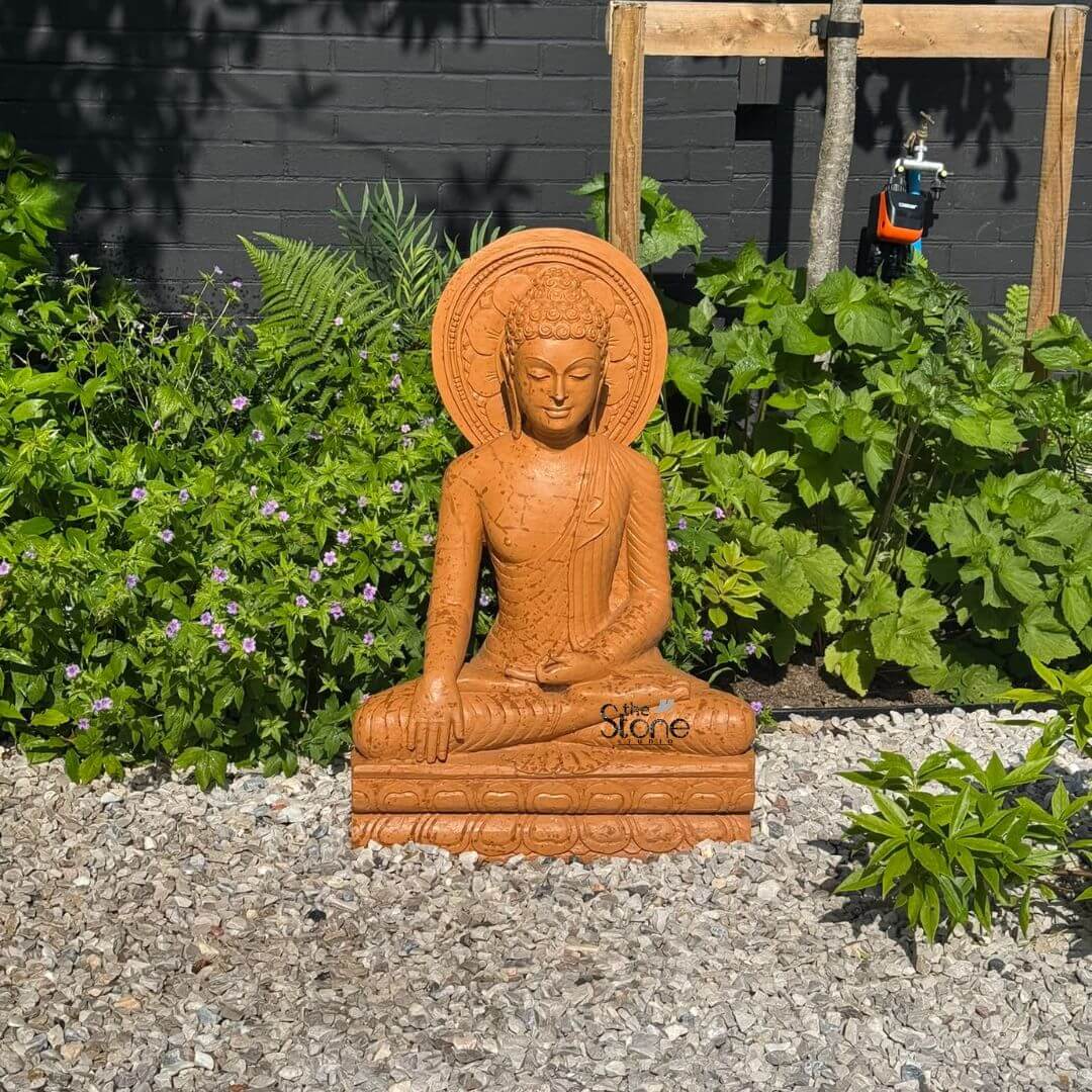 3 feet Bhumisparsha Buddha Statue 6 A serene 3-foot Buddha statue crafted from warm-toned sandstone sits gracefully in a contemplative pose within a verdant garden setting likely in Walton on Thames London as suggested by its article context. The Buddha is depicted with closed eyes a gentle smile and elongated earlobes conveying a sense of profound peace and inner tranquility. His hands rest in his lap in the Dhyana Mudra a gesture of meditation and focus which is characteristic of statues designed for calming environments. The natural texture of the sandstone is visible offering an earthy and grounded feel to the sculpture perfect for an outdoor spiritual space. The statue is positioned on a rectangular base also made of sandstone which rests on a bed of light-colored gravel. Surrounding the Buddha is a lush array of green plants including ferns flowering ground cover with small purple blossoms and larger leafy bushes creating a natural and harmonious backdrop. In the background a dark fence and the base of a tree are visible along with what appears to be a small outdoor spigot or watering device indicating a residential or cultivated garden environment typical of areas like Walton on Thames. The interplay of light and shadow subtly highlights the contours of the Buddha's face and robes, enhancing its spiritual presence. This 3-foot sandstone Buddha statue serves as a focal point radiating calm amidst the natural growth of the garden. Its size and material make it a suitable piece for a personal meditation space or a decorative element in a garden in London aligning perfectly with an article discussing Buddha Statue in Walton on Thames emphasizing both its artistic craftsmanship and its potential role in fostering tranquility within an urban or suburban landscape.