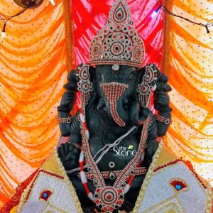 A 15-inch Ganesha statue carved from green stone is adorned magnificently evoking the grandeur of a Dagdusheth Ganpati Stone Statue in Mumbai. The statue depicts Lord Ganesha in a seated posture his elephant head crowned with an elaborate sparkling diadem. This crown along with his ears and trunk is lavishly embellished with red white, and gold beadwork and intricate designs radiating opulence and divine majesty. The deep green hue of the stone is visible beneath the adornments adding a rich natural foundation to the vibrant decorations. Ganesha's body is further decorated with a vibrant red white and gold beaded garland that drapes across his chest emphasizing his sacred presence. His arms holding traditional attributes are also adorned with matching bejeweled armlets. The statue is seated on a base draped with ornate fabric in white and gold featuring pearl-like detailing and red accents resembling ceremonial attire. The meticulous craftsmanship of both the stone carving and the elaborate embellishments speaks to a high level of artistry and devotion. The backdrop for this divine display consists of rich flowing orange and red fabric drapes illuminated by warm string lights creating a festive and reverent atmosphere characteristic of a major temple or celebration like those in Mumbai. The overall presentation is one of profound beauty and spiritual reverence making this 15-inch green stone Ganesha statue a captivating representation for an article about the revered Dagdusheth Ganpati.