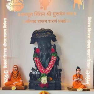 A beautifully arranged home altar or shrine features a central dark stone statue of Lord Dattatreya Dutta Bhagwan flanked by two smaller statues of revered sages all against a bright illuminated background with golden Sanskrit script. The central Dattatreya statue carved from deep black stone depicts the deity with three heads representing Brahma Vishnu and Shiva each with serene expressions. He is adorned with fresh vibrant floral garlands prominently a thick U-shaped garland of red and white flowers likely roses and jasmine providing a striking contrast against the dark stone. Additional red roses and green foliage are tucked into the statue enhancing its devotional presentation. The statue is placed on an intricately carved dark base signifying its importance. Flanking the Dattatreya statue on either side are two smaller statues of sages or spiritual teachers both seated in meditative postures and wearing bright orange robes. The sage on the left has a calm benevolent expression with hands in a gesture of blessing or teaching. The sage on the right, equally serene has a topknot and hands crossed exuding wisdom. These figures are placed on light-colored wooden bases harmonizing with the overall spiritual display. Small yellow and white flowers are placed at the feet of these flanking statues indicating offerings and reverence. The background is a well-lit smooth white wall adorned with elegant golden Sanskrit calligraphy adding a sacred and ethereal ambiance. The script appears to be devotional verses or mantras further reinforcing the spiritual context of the display. Decorative golden lotus motifs and possibly images of other deities are subtly integrated into the upper corners of the background. On the vertical edges intricate white cut-out patterns possibly resembling flames or traditional designs add an artistic border to the shrine. This entire setup creates a profound and visually appealing devotional space ideally suited for an article about Dutta Bhagwan statues in areas like Chembur highlighting the cultural richness and spiritual significance of such idols in Hindu worship.