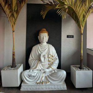 A majestic 5-foot FRP Fiber Reinforced Polymer Buddha statue is prominently displayed suggesting its presence in Durg embodying serenity and modern elegance. The Buddha is depicted in a meditative pose seated in full lotus position with his hands positioned in the Vitarka Mudra symbolizing teaching and intellectual discussion. His robe is a pristine white with intricate golden accents that highlight the graceful folds of the fabric adding a luxurious touch to the minimalist design. The Buddha's face exudes profound peace with gently closed eyes and a subtle smile while his hair is rendered in a textured golden finish culminating in a prominent topknot. The statue's smooth white finish and golden detailing create a striking contrast enhancing its spiritual presence and aesthetic appeal. The use of FRP ensures a durable yet relatively lightweight statue suitable for various indoor and outdoor settings which is a practical consideration for a piece of this size. The craftsmanship is evident in the refined features and the flowing lines of the robe contributing to its overall artistic quality and ability to evoke tranquility. The Buddha statue rests on a matching white base also adorned with golden accents providing a stable and harmonious foundation. It is flanked by two tall elegant potted palm trees with verdant fronds planted in square white planters filled with white pebbles. The backdrop is a clean dark wall which further emphasizes the statue's bright appearance. This composition creates a balanced and serene environment making this 5-foot FRP Buddha statue a captivating subject for an article exploring such statues and their role in contemporary decor especially in places like Durg.