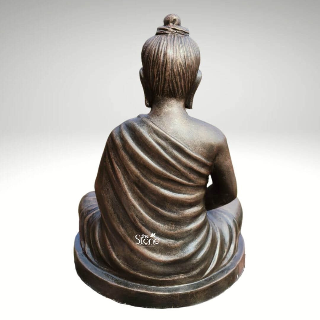 2 Feet Fibre Buddha Statue 5 A serene 2-foot fiber Buddha statue finished with a metallic bronze-like coating is gracefully presented from a rear view against a neutral background making it an ideal piece for home decor. The Buddha is depicted in the classic Dhyana Mudra seated in full lotus position with his hands resting gently in his lap symbolizing deep contemplation and enlightenment. From this perspective the prominent topknot of his hair is clearly visible along with the dignified posture of his head and back conveying profound inner peace and spiritual wisdom. The lustrous finish of the fiber material made to resemble aged bronze adds a sense of timeless dignity and elegance to the sculpture. The detailing on the idol is meticulously rendered with particular emphasis on the graceful natural folds of his monastic robe that drape softly over his back and shoulders extending down to his crossed legs. The light plays across the contours of the statue highlighting the subtle texture of the robe and the smooth surface of the fiber. The bronze-like appearance effectively mimics the rich patina of an antique metal piece offering both aesthetic appeal and the practical benefits of durable fiber construction which is lighter yet robust making it suitable for various home decor settings. The Buddha idol rests on a simple low and subtly curved rectangular base which seamlessly integrates with the overall form providing stability without distracting from the main figure. This 2-foot fiber Buddha statue with its elegant bronze-like finish and peaceful demeanor embodies tranquility and spiritual grace serving as a compelling subject for an article exploring aesthetically pleasing and durable Buddha statues suitable for enhancing the serene ambiance of any home.