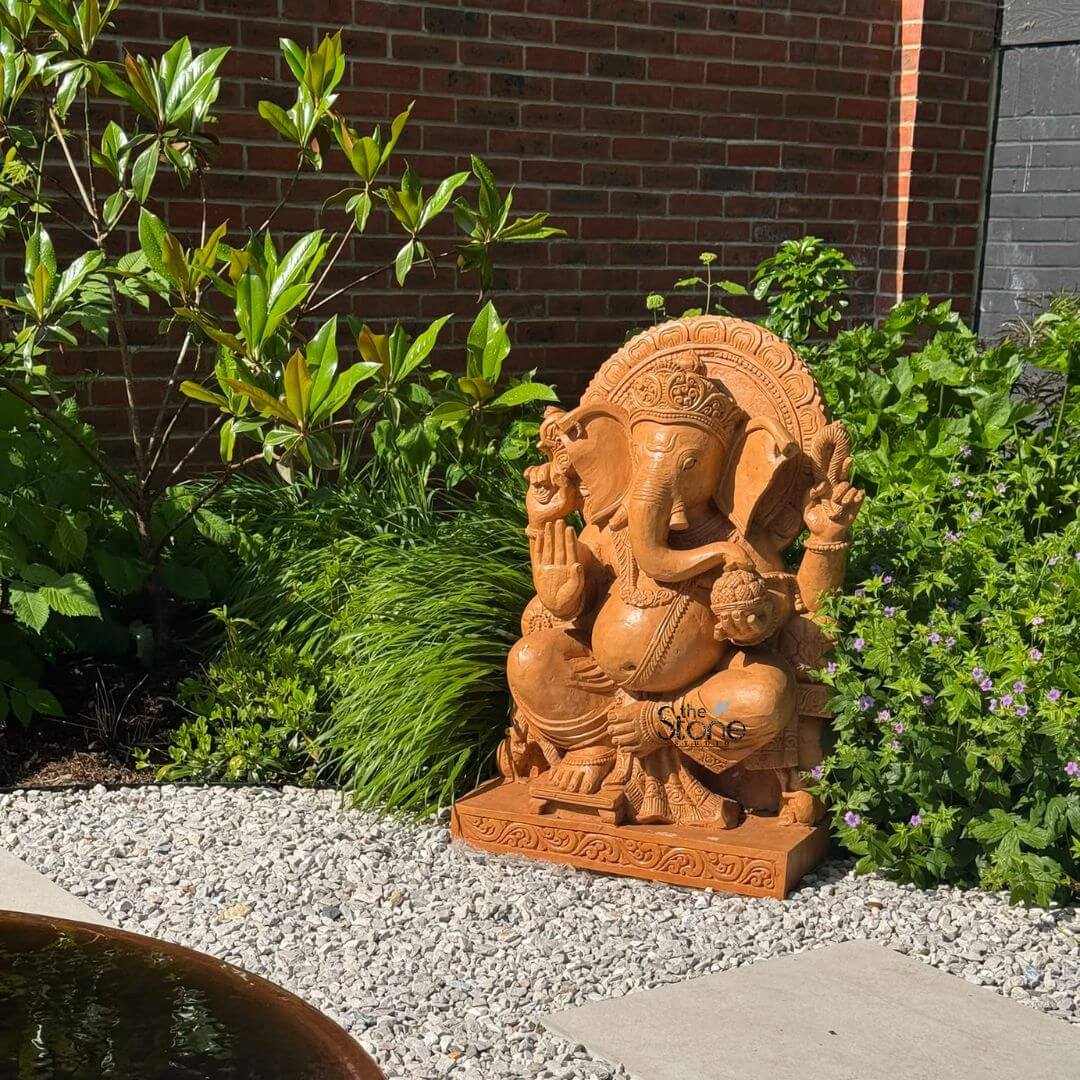 Ganpati Idol 3ft 5 A charming 3-foot Ganesha statue crafted from rich reddish-brown sandstone is beautifully situated in an outdoor garden setting evocative of a tranquil space in Walton on Thames London. Lord Ganesha is depicted in a seated relaxed posture embodying wisdom and auspiciousness. His right hand is raised in a gesture of blessing while his left hand gently holds a modak signifying prosperity and good fortune. The intricate carvings on his crown jewelry and pedestal are finely detailed showcasing the craftsmanship of the sandstone material. The statue's warm tone contrasts appealingly with the surrounding greenery and natural light. The Ganesha statue rests on a decorative carved sandstone base placed on a clean bed of light-colored gravel. This meticulously maintained area is bordered by a paved walkway in the foreground and lush garden beds in the background. The garden features a variety of plants including tall grasses leafy shrubs and flowering ground cover with small purple blooms. Behind the foliage a warm-toned brick wall is visible on the left and a darker wall on the right suggesting a typical residential garden or courtyard in an urban or suburban area like Walton on Thames. In the lower left foreground a glimpse of a terracotta-colored water feature or planter adds another element of natural beauty to the scene. The overall composition creates a harmonious blend of spiritual artistry and natural landscape. This 3-foot sandstone Ganesha statue with its inviting presence and detailed design is an ideal subject for an article discussing Ganesha Statue in Walton on Thames London highlighting its aesthetic appeal as a garden focal point and its cultural significance within a contemporary British setting.