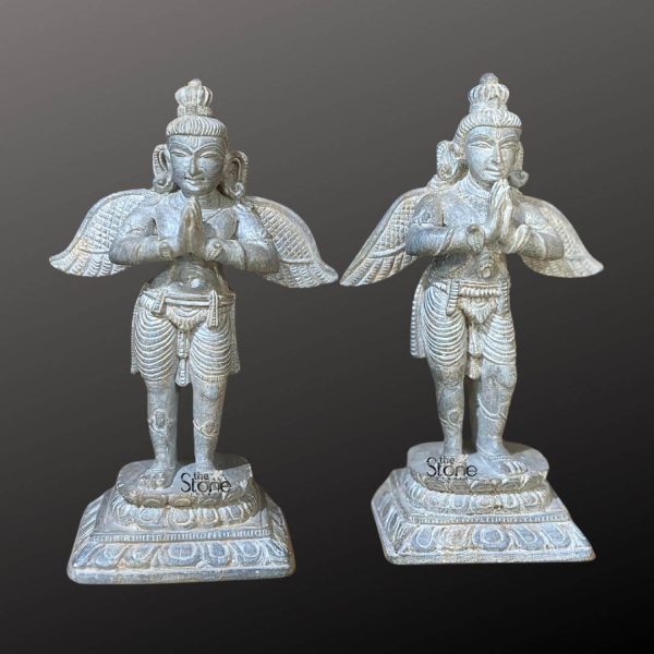 A magnificent pair of 6-inch Garuda statues carved from green stone stands majestically against a dark neutral background highlighting their intricate details and divine presence. Each statue depicts Garuda the mythical bird-like creature and vahana of Lord Vishnu in a reverent standing pose with hands pressed together in Anjali Mudra. The greenish hue of the stone with its subtle variations gives the statues an aged, weathered and naturalistic appearance emphasizing their traditional craftsmanship. Both Garuda figures are identically sculpted showcasing finely detailed features including their crowns expressive faces and powerful outstretched wings. The wings, a prominent feature are meticulously carved with individual feathers conveying a sense of readiness for flight and immense strength. Their lower bodies are adorned with intricately detailed dhotis or skirts adding to their divine warrior aesthetic. Each statue stands on a square multi-tiered pedestal also intricately carved with traditional patterns providing a stable and ornate base that enhances their grandeur. This image is ideally suited for an article discussing Garuda statue pairs for temple decor underscoring their significance as guardians and symbols of devotion. The 6-inch size makes them suitable for various temple niches or home altars while the durable green stone ensures their longevity. Presented as a pair they embody spiritual symmetry and represent the faithful attendance and power associated with Garuda making them perfect for enhancing the sacred ambiance of any devotional space.