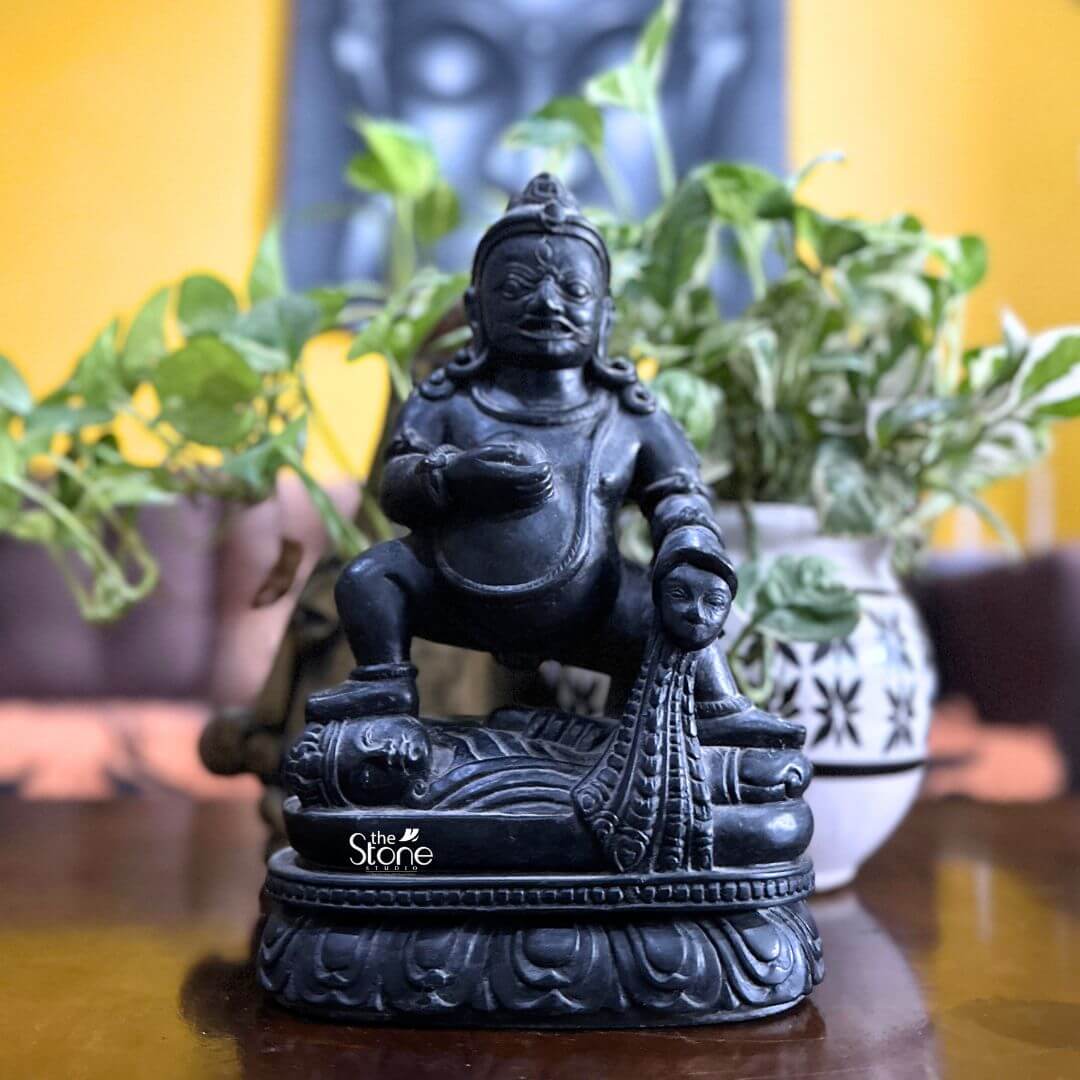 Black Stone Jambhala Statue 9" 3 A striking 9-inch Jambhala statue meticulously carved from black stone is prominently displayed on a polished wooden surface likely a table in an inviting indoor setting. The deity is depicted in a dynamic squatting posture exuding an aura of robust strength protection and generosity. Jambhala's facial features are distinct with wide intense eyes a prominent mustache and a stern yet benevolent expression conveying his role as a guardian of wealth and dispeller of poverty. The deep polished black stone exquisitely captures the statue's muscular physique the folds of his belly and the rich textures of his attire and ornamentation. In his left hand Jambhala holds a spherical object traditionally identified as a jewel or a fruit representing the bestowal of riches and spiritual abundance. His right hand rests upon a stylized mongoose a significant attribute in Buddhist iconography shown gracefully expelling a cascade of jewels symbolizing his power to grant material wealth. The craftsmanship extends to the intricate details of his crown earrings necklaces and armlets all precisely rendered in the dark stone. The statue is seated on a beautifully sculpted lotus pedestal a profound symbol of purity and spiritual growth providing a sacred foundation. In the soft-focus background green potted plants and a large artistic print or mural on a warm yellow wall add to the vibrant spiritual ambiance of the room. This image is ideally suited for an article focused on Jambhala statues for wealth and prosperity showcasing a prime 9-inch black stone example that embodies both spiritual significance and artistic mastery within a living space. The black stone material gives the statue a timeless and authoritative presence making it a compelling piece for a home altar office or personal collection where the intention is to invite good fortune and abundance. The detailed depiction of Jambhala's attributes particularly the jewel-spitting mongoose directly communicates his benevolent function in Buddhist practice making this a powerful visual for the article's discussion on this revered deity of wealth and prosperity.