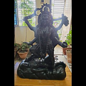 A powerful and imposing 3-foot Kali Maa Stone Statue carved from blackstone is dynamically displayed in what appears to be an indoor setting with natural light possibly a studio or a home with large windows. The multi-armed goddess stands triumphantly over the prostrate body of Lord Shiva a classic depiction of her ultimate power and transcendental nature. Kali Maa is depicted with a fierce yet serene expression her multiple arms holding various implements including a sword a severed head and a bowl symbolizing the destruction of evil liberation and sustenance. Her matted hair flows freely and she is adorned with garlands of skulls or beads, armlets, and a crown. The blackstone material gives the statue a commanding and ancient presence highlighting the intricate details of her iconography and the depth of the carving. The base of the statue is a complex composition depicting the prostrate form of Lord Shiva beneath Kali Maa's feet. Shiva is serene accepting her dance of cosmic dissolution symbolizing the triumph of Shakti over inert consciousness. The dark blackstone allows for deep carving, which accentuates the details of both figures from Kali Maa's powerful stance and flowing garments to Shiva's calm visage. The statue is placed on a light-colored wooden surface which provides a simple yet effective contrast making the dark sculpture stand out and emphasizing its central importance. The background shows large windows allowing ample natural light to illuminate the scene with potted plants visible outside creating a subtle green backdrop. This juxtaposition of the fierce deity with a serene natural environment suggests a harmonious integration of spiritual power within a living space. The entire setup emphasizes the statue's artistic merit profound spiritual significance and its awe-inspiring presence making it an excellent visual for an article about Kali Maa Stone Statues particularly those crafted from blackstone and revered in places like Kolkata showcasing their ability to command attention and inspire devotion.