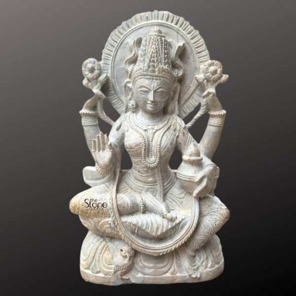A resplendent 12-inch Lakshmi idol meticulously carved from green stone is presented against a dark neutral background emphasizing her divine beauty and intricate details. The goddess of wealth and prosperity is depicted in a traditional seated posture likely Padmasana emanating grace and benevolence. The greenish hue of the stone with its subtle variations gives the statue an aged timeless appearance highlighting its traditional craftsmanship and serene presence making it ideal for a puja room. Lakshmi is shown with four arms holding traditional attributes that symbolize her divine powers. In her upper hands she gracefully holds two lotus flowers representing purity beauty and spiritual growth. One of her lower hands is raised in Abhaya Mudra while the other holds a kalasha or a similar auspicious vessel. Her face is serene and benevolent adorned with a regal crown and intricate jewelry including necklaces armlets and bangles all finely sculpted. A circular halo, or prabhamandala frames her head featuring radiating lines that suggest divine light and energy. The base of the statue is carved with delicate lotus petals further reinforcing her association with purity and auspiciousness. A small owl her traditional vahana is subtly depicted near her feet. This image is ideally suited for an article discussing Lakshmi idols for a puja room showcasing a prime 12-inch green stone example that embodies both spiritual significance and artistic mastery. The durable green stone ensures its longevity and aesthetic appeal. The detailed depiction of Lakshmi's attributes graceful posture and serene expression directly conveys her benevolent function as the bestower of wealth, prosperity and spiritual well-being, making it a compelling visual for the article's focus on creating a sacred and abundant space for worship.