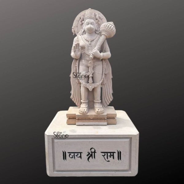 A majestic 7-foot Large Hanuman Statue meticulously carved from sandstone stands prominently on a multi-tiered pedestal radiating power and devotion. Hanuman is depicted in a standing posture with one hand raised in a gesture of blessing symbolizing reassurance and protection while his other hand firmly grasps his formidable mace which rests on his shoulder. His powerful muscular physique is evident adorned with traditional ornaments including necklaces armlets and a sacred thread. His face features a serene yet determined expression with his characteristic monkey-like features a beard and a crown. The sandstone material gives the statue a natural earthy tone with subtle variations enhancing its antique and spiritual appeal. Behind Hanuman's head a beautifully carved prabhavali frames his divine presence featuring intricate patterns that add to the grandeur of the sculpture. His attire includes a finely draped dhoti or lower garment with flowing pleats that suggest movement and grace complementing his powerful stance. The details of his feet resting firmly on a small carved sandstone base are also visible further grounding the powerful figure. The craftsmanship highlights deep relief carvings and a polished yet natural finish characteristic of high-quality sandstone artistry. The entire Hanuman statue rests on a substantial multi-tiered white or light-colored pedestal. The front face of this lower pedestal features a prominent inscription in Devanagari script which reads ॥ जय श्री राम ॥ Jaya Shri Rama signifying Victory to Lord Rama a direct reference to Hanuman's ultimate devotion and his role in the Ramayana. The dark, gradient background effectively isolates the statue and its pedestal drawing full attention to its imposing size intricate details and spiritual message. This image serves as an excellent visual for an article about Large Hanuman Statues particularly those crafted from sandstone emphasizing its artistic merit devotional significance and its impressive presence as a revered deity.