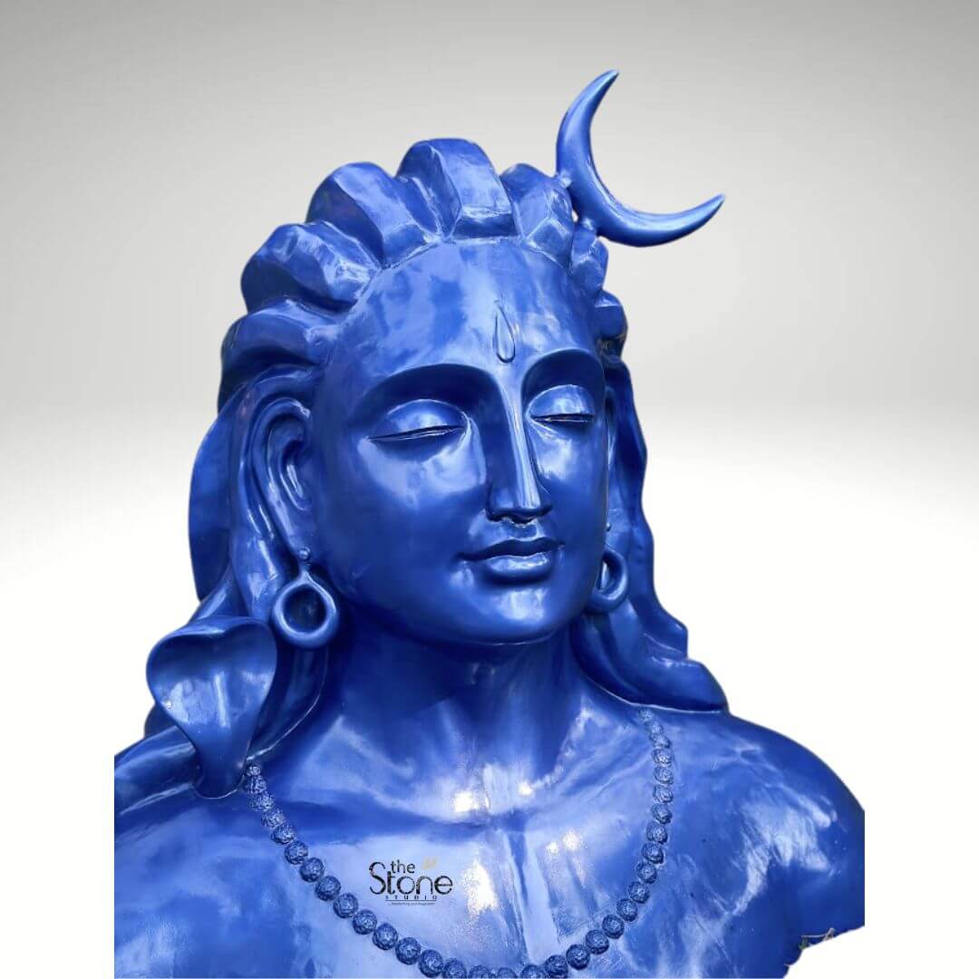 Blue Adiyogi Shiva Statue 4ft 8 A striking 4-foot fiber statue of the Lord Shiva Adiyogi bust is prominently displayed against a plain background serving as a powerful visual for an article about such revered sculptures. The sculpture captures the upper torso and head of Lord Shiva in his Adiyogi form depicted with closed eyes in deep meditation conveying profound inner peace and wisdom. His iconic third eye is subtly marked on his forehead and a prominent crescent moon gracefully adorns his matted locks which are styled upwards and flow around his shoulders symbolizing his ascetic nature. The entire statue is rendered in a vibrant deep blue color characteristic of Shiva's divine complexion. Shiva's ears are adorned with traditional large ring earrings and a detailed serpent is subtly carved around his neck along with a beaded necklace adding to his divine iconography. The intricate detailing on his hair ornaments and facial features showcases remarkable artistry. The smooth glossy finish of the fiber material enhances the statue's vibrant blue hue making it a visually captivating piece that evokes both ancient reverence and modern artistic interpretation. The robust fiber construction ensures the statue is durable yet relatively lightweight suitable for various indoor and outdoor settings. The statue's bust-like form, with its broad shoulders and serene countenance exudes a powerful yet tranquil presence making it a compelling focal point. This 4-foot fiber Lord Shiva Adiyogi statue truly embodies spiritual power and artistic grace, with its intense blue color adding to its mystique. Its commanding yet peaceful presence makes it a powerful symbol of yoga and ultimate liberation perfect for inspiring contemplation and reverence.
