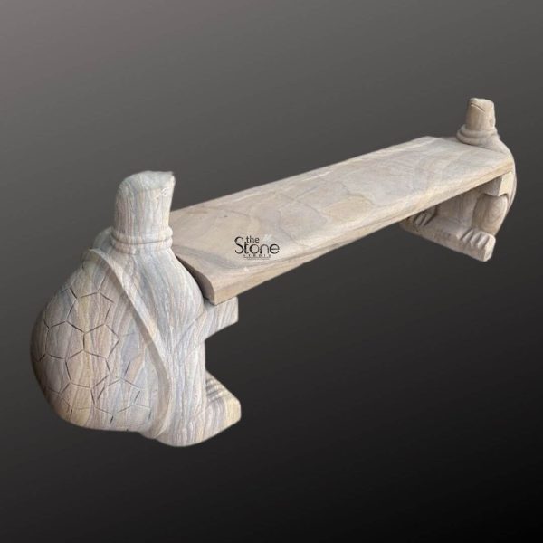 A unique Outdoor Garden Stone Bench intricately carved from light-colored sandstone is presented against a dark gradient background emphasizing its sculptural qualities. The bench is composed of a thick rectangular slab forming the seating surface supported at each end by exquisitely sculpted animal figures possibly stylized turtles or similar mythical creatures acting as decorative and structural legs. The sandstone has a natural somewhat rustic finish with visible grain and subtle variations in tone indicating its organic origin and handcrafted nature. The supporting figures at each end of the bench are highly detailed. One figure prominently visible on the left features a rounded shell-like back with a geometric pattern resembling scales or segments leading to a gracefully carved neck and head. The texture and lines on the stone evoke a sense of ancient craftsmanship. The overall design suggests a blend of artistic flair and functional stability making it not just a piece of furniture but a decorative garden element. The other supporting figure partially visible on the right mirrors the design of the first ensuring symmetry and balance. The angle of the photograph highlights the three-dimensionality of the bench showcasing the depth of the carving and the robust construction. The dark background helps to isolate the bench drawing attention to its light color and the fine details of its stone texture. This image effectively illustrates an article about Outdoor Garden Stone Benches particularly those made from sandstone emphasizing their durability artistic design and their ability to add a touch of elegance antiquity and unique charm to any outdoor living space or garden landscape.