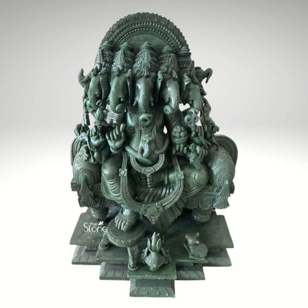 A magnificent 3-foot Panchamukhi Ganesha statue meticulously carved from green stone stands prominently against a clean white background emphasizing its grandeur and intricate details. Lord Ganesha is depicted in his powerful five-headed form with the central head facing forward and four other heads radiating around it signifying omnipresence and the ability to bless from all directions. The deep green hue of the stone with its subtle variations gives the statue a rich, aged and traditional appearance highlighting the master craftsmanship involved in this complex carving. The statue features a seated Ganesha with numerous arms each holding various traditional attributes such as a modak a goad a noose and other symbolic items representing his divine powers and functions. The intricate details of his crowns, large ears trunks, and extensive ornamentation including necklaces and armlets are exquisitely rendered. A grand ornate halo frames the five heads featuring detailed carvings that enhance the divine aura of the deity. The figure is seated on an elaborately sculpted throne or pedestal which is further supported by a multi-tiered stepped base all carved from the same green stone adding to its imposing presence. A small mouse Ganesha's traditional vahana is subtly depicted near his feet. This image is ideally suited for an article discussing Panchamukhi Ganesha statues showcasing a prime 3-foot green stone example that embodies immense spiritual significance and artistic mastery. The substantial size and durable green stone material make it an impressive and lasting addition to any temple large home altar or prominent spiritual space. The detailed depiction of Ganesha's five heads and numerous attributes directly conveys his benevolent function as the remover of obstacles bestower of success and guardian of prosperity from all directions making it a powerful visual for the article's focus on this revered form of the deity.