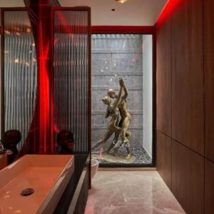 This image showcases a striking contemporary interior design with a focus on an artistic Pop Art Statue set within a sophisticated and mood-lit space. The central feature is a large bronze-toned statue depicting two figures in a dynamic intertwined dance pose conveying movement and emotion. The statue possibly made of metal or a metallic-finished material stands on a bed of dark pebbles visible through a large floor-to-ceiling window or glass partition. This window opens into what appears to be an enclosed outdoor or garden area with a textured grey stone wall, creating a dramatic backdrop for the sculpture. The interior space exudes a modern and luxurious ambiance characterized by rich materials and dramatic lighting. On the left a sleek vanity area features a wide rectangular white basin and a large mirror with a ribbed or fluted glass panel beside it reflecting warm red light that bathes the space in a vibrant glow. This red lighting is also subtly visible along the ceiling creating an inviting yet bold atmosphere. The walls on the right are paneled with warm reddish-brown wood adding texture and sophistication. The floor is made of smooth light-colored marble or polished stone tiles reflecting the ambient light and contributing to the spacious feel. The overall design suggests a high-end bathroom or dressing area that seamlessly integrates art and modern aesthetics. The statue with its contemporary style and emotional intensity serves as a powerful focal point illustrating an article about Pop Art Statues particularly those found in urban centers like Delhi and their role in enhancing modern interior and exterior spaces.