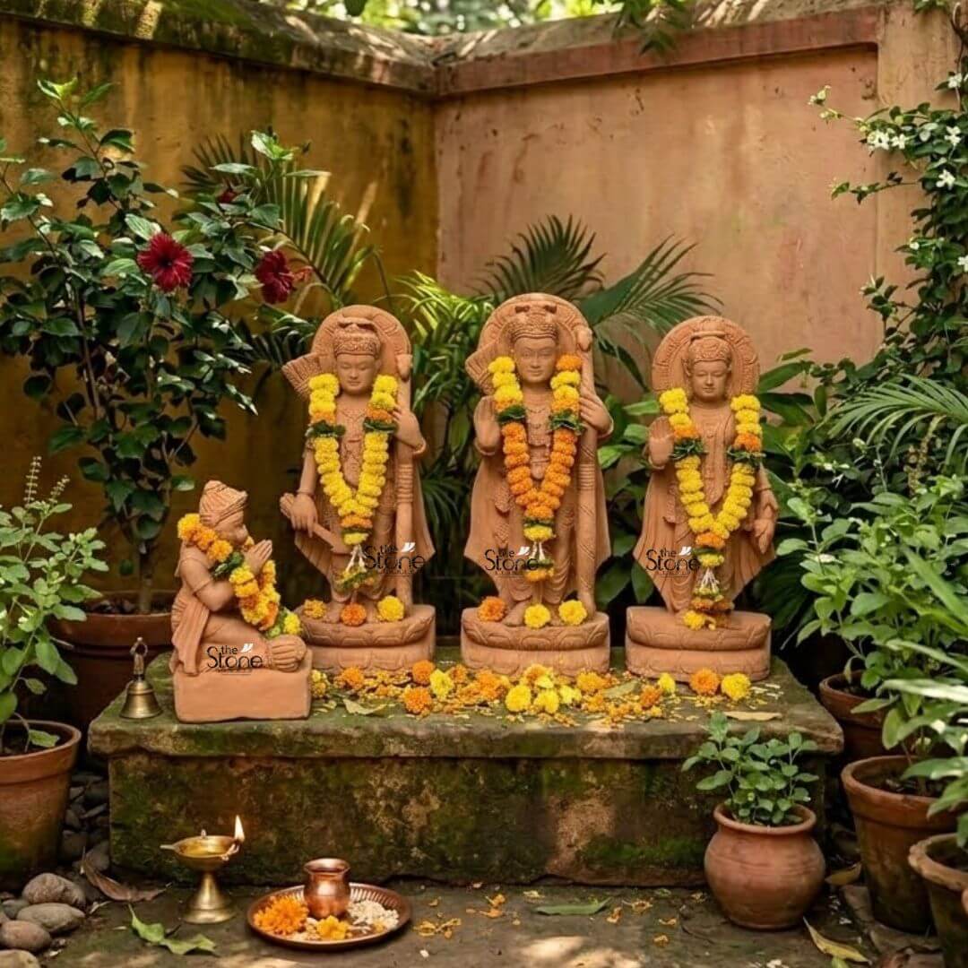 Ram Darbar Idol for Home Temple 2ft 3 A beautifully arranged 2-foot sandstone Ram Darbar idol set is reverently displayed in a lush outdoor garden setting ideal for an article on Ram Darbar idols in stone. The set comprises four figures Lord Rama Lakshmana Sita and a kneeling Hanuman collectively forming a revered divine assembly. Lord Rama stands prominently in the center holding his bow and arrow embodying righteousness and valor. To his left stands Lakshmana also armed and to his right the graceful Sita with her hand raised in blessing. Each of these standing figures is adorned with elaborate crowns intricate jewelry and flowing traditional garments, all meticulously carved from the warm earthy sandstone. Vibrant yellow and orange marigold garlands adorn each deity symbolizing devotion and festivity. Behind the heads of Rama Lakshmana, and Sita circular halo-like structures are intricately carved with traditional patterns further accentuating their divine aura. The craftsmanship is evident in every detail from the serene expressions on their faces to the delicate folds of their attire and the intricate patterns of their ornaments. The sandstone's natural texture and warm hue give the statues an authentic, ancient feel enriching their spiritual presence and highlighting the artisans' skill blending seamlessly with the natural environment. In the foreground kneeling respectfully is Hanuman carved in a posture of profound devotion with his hands clasped in reverence. The entire assembly rests on a moss-covered stone platform surrounded by potted plants and scattered marigold petals. A small brass bell, a lit diya and offerings of flowers and sweets are placed before the deities creating an atmosphere of puja. The warm, dappled sunlight illuminates the scene making this 2-foot sandstone Ram Darbar idol set a captivating representation of spiritual devotion and artistic beauty for any home or garden.