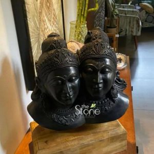 An 18-inch Shiva Parvati Blackstone statue is prominently displayed showcasing the divine couple side-by-side carved from a deep lustrous black stone. The statue captures the busts of Lord Shiva and Goddess Parvati with Shiva on the left and Parvati on the right their faces exuding serene expressions of grace and devotion. Shiva is depicted with his characteristic matted locks styled into a topknot adorned with intricate details and possibly a crescent moon while Parvati's headpiece is equally elaborate suggesting a crown or ornate hair arrangement. Both figures possess delicate facial features including almond-shaped eyes well-defined noses and gentle smiles conveying their divine presence. The blackstone material lends a striking contrast to any setting absorbing light and highlighting the sculptural forms and intricate carvings. The details extend beyond their faces to their necks and shoulders where subtle ornamentation like necklaces or garment folds are suggested adding to the richness of the depiction. The craftsmanship is evident in the smooth finish of the stone and the precision of the carving which brings out the traditional iconography associated with these Hindu deities. The statue is positioned on a light-colored wooden surface which provides a natural and warm base that beautifully offsets the dark stone. The background of the image features a softly blurred interior setting suggesting a home or studio environment. Hints of other decorative elements such as artwork on a wall vertical plants and patterned textiles can be discerned providing context to how such a statue might be integrated into a living space. This image effectively illustrates an article about Shiva Parvati stone decor statues particularly those crafted from blackstone and available in places like Ernakulam emphasizing their cultural significance artistic beauty and their role as elegant and spiritual home decor pieces.
