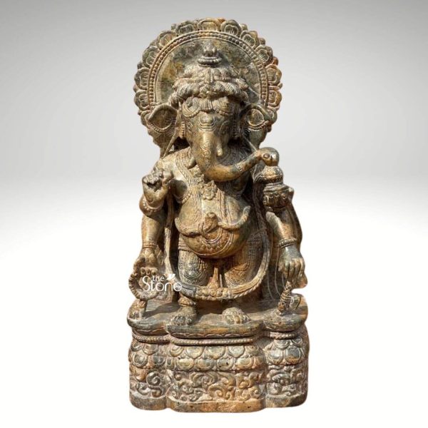 A beautifully carved 12-inch Standing Lord Ganesha Murti crafted from green stone is presented against a plain light grey background allowing its intricate details to stand out. Ganesha is depicted in a dynamic standing pose with a slight bend in his knees conveying a sense of gentle movement and divine presence. His iconic elephant head with large ears a prominent trunk curled towards the left and one broken tusk is central to his identity as the remover of obstacles. His multiple arms hold various attributes one hand gracefully holds a modak while another may hold an axe or a lotus symbolizing wisdom prosperity and spiritual liberation. The green stone material gives the statue a rich earthy and slightly weathered appearance highlighting the deep relief carvings and textures. Ganesha is adorned with traditional ornaments including necklaces armlets and a sacred thread across his chest all meticulously detailed. A patterned dhoti or lower garment is also visible flowing naturally with his stance. Behind his head an elaborately carved prabhavali with concentric floral or geometric patterns frames his divine countenance adding to the grandeur of the sculpture. The intricate carvings extend to his robust belly a symbol of prosperity. The Murti stands on a multi-tiered ornate base that is integrated with the main sculpture. The base itself is richly carved with traditional motifs possibly lotus petals or other auspicious designs providing a stable and aesthetically pleasing foundation. The overall craftsmanship reflects a deep understanding of traditional Indian sculptural art making it an ideal piece for an article about Standing Lord Ganesha Murtis particularly those made from green stone. The natural color and detailed carving make it a significant object of devotion and a beautiful addition to any spiritual or decorative setting.