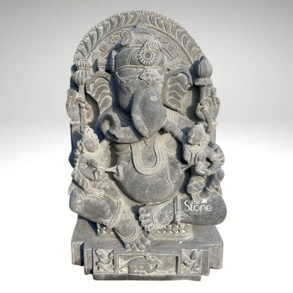 A magnificent black stone Ganesha statue depicting Lord Ganesha flanked by his consorts Riddhi and Siddhi is prominently featured against a clean neutral background, emphasizing its intricate details and sacred presence. This exquisite sculpture ideal for an article about Ganesha with Riddhi Siddhi statues showcases the deities with profound artistic and spiritual qualities. Lord Ganesha is seated centrally in a traditional posture radiating an aura of wisdom, prosperity and auspiciousness. His trunk elegantly curls to his left touching a modak in his lower left hand symbolizing abundance. His other hands hold traditional attributes including a pasha an ankusha and his broken tusk. Ganesha is richly adorned with meticulously carved traditional jewelry including a regal crown elaborate necklaces armlets and anklets all integrated seamlessly into the dark black stone. His large fanned ears and expressive eyes beneath arched brows convey a sense of calm and deep insight. His prominent belly a hallmark of Ganesha, symbolizes prosperity and the capacity to encompass all of existence. Flanking Ganesha are the smaller figures of Riddhi and Siddhi seated gracefully on either side. Riddhi is typically seen on Ganesha's right and Siddhi on his left each holding symbolic items and adorned with their own intricate jewelry reflecting their divine roles. The entire ensemble is set within an intricately carved prabhavali that frames the deities adorned with traditional motifs further enhancing the divine composition. The base of the statue is also highly detailed featuring a row of smaller figures including Ganesha's vahana the mouse adding narrative depth. The craftsmanship of the black stone sculpture is exceptional with its smooth dark finish accentuating the fine details and robust forms of the deities. The inherent qualities of the stone give the statue a timeless and dignified presence suitable for a temple home temple or a prominent spiritual space. This Ganesha with Riddhi Siddhi statue is not merely a decorative item but a powerful spiritual artifact inspiring devotion and bringing blessings of success prosperity and spiritual fulfillment.