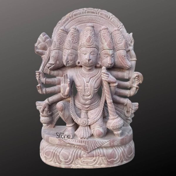 A magnificent 12-inch Panchamukhi Hanuman statue carved from pinkstone is prominently displayed against a dark gradient background making it an excellent visual for an article about Stone Panchamukhi Hanuman statues. The statue depicts Lord Hanuman in his powerful five-headed form with his central monkey face flanked by four other faces of divine beings. He is sculpted in a dynamic kneeling posture one knee raised signifying readiness for action and unwavering devotion. His multiple arms extend outwards each holding a traditional weapon or symbol including a prominent Gada in one of his lower right hands symbolizing immense strength. The statue is richly adorned with intricate jewelry including elaborate necklaces armlets and a detailed crown on each head. The fine details of the carving are evident in the expressive faces the flowing lines of his dhoti and the powerful musculature all brought to life by the warm subtle pink hue of the stone. Behind the heads a circular halo-like structure is intricately carved with traditional patterns further accentuating his divine aura and emphasizing his formidable presence. The craftsmanship showcases remarkable artistry capturing the fierce yet benevolent nature of Lord Hanuman. Panchamukhi Hanuman rests on a multi-layered intricately carved lotus pedestal also made from pinkstone providing a stable and aesthetically pleasing foundation with traditional motifs. The natural texture and color of the pinkstone highlight the deep textures and intricate patterns of the carving making every detail stand out. This 12-inch pinkstone Panchamukhi Hanuman statue truly embodies spiritual power and artistic excellence serving as a powerful and beautiful representation of the beloved deity.