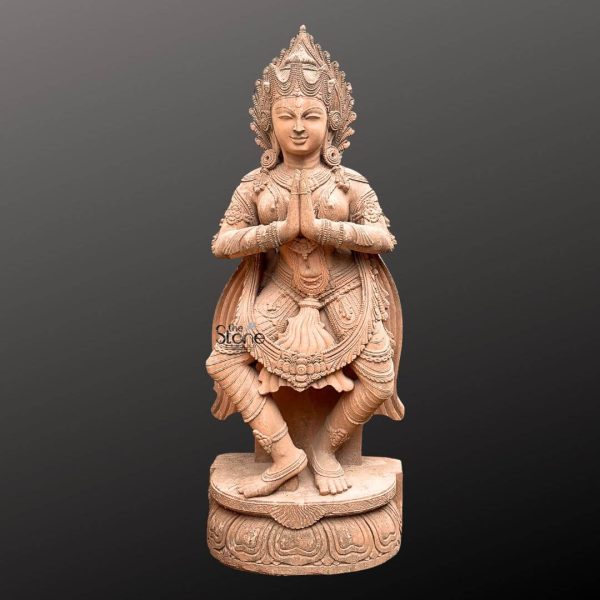 A magnificent 6-foot Traditional Indian Devdasi Sculpture meticulously carved from brown sandstone stands gracefully against a dark gradient background emphasizing its artistic and cultural significance. The sculpture depicts a Devdasi a female temple dancer in a kneeling or half-seated posture with hands pressed together in a respectful gesture of Namaste or prayer. Her face exudes a serene and welcoming expression adorned with an elaborate crown or headdress that features intricate carvings signifying her revered status. The sandstone material gives the statue a warm earthy brown tone highlighting the fine details of the carving and its antique appeal. The Devdasi is richly adorned with traditional Indian jewelry including multiple necklaces armlets wristlets and anklets all intricately sculpted into the stone. Her attire possibly a flowing sari or traditional dancer's costume is rendered with exquisite detail showing drapes and folds that accentuate her graceful posture. Her waist is cinched with an ornate belt and tassels or decorative elements hang from her waist. The sculptural artistry is evident in the delicate features of her face, her slender fingers and the overall fluidity of her form conveying both devotion and artistic grace. The detailed carvings on her costume reflect classical Indian temple art. The sculpture rests on a multi-tiered integrated lotus base also carved from brown sandstone featuring detailed lotus petals that provide a beautiful and symbolic foundation. The intricate craftsmanship evident throughout the sculpture from the expression on her face to the details of her adornments and the ornate base makes this an ideal representation for an article about Traditional Indian Devdasi Sculptures. Its imposing size and authentic traditional design make it a significant artistic and cultural object evoking a sense of ancient Indian heritage, devotion and the beauty of classical dance forms.