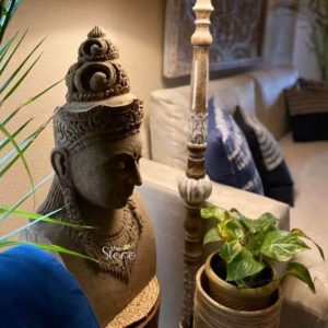 A magnificent 2-foot Vishnu Stone Head Statue carved from antique-look sandstone is beautifully displayed in a warmly lit interior setting exuding a serene and spiritual ambiance. The statue focuses on the head and upper torso of Lord Vishnu intricately sculpted with a majestic multi-tiered crown adorned with traditional motifs. Vishnu's face bears a calm and benevolent expression with finely carved features including half-closed eyes a gentle smile and a defined nose conveying profound peace and divinity. His elongated earlobes are also visible a common characteristic in sacred sculptures. The sandstone material has a rich earthy brown tone enhanced by an antique finish that gives it a timeless weathered appearance. The statue's upper chest is adorned with an elaborate necklace detailed with traditional patterns and a sacred thread is subtly indicated enhancing its regal and divine portrayal. The texture of the sandstone is visibly rich showcasing the hand-carved artistry and the natural variations within the stone contributing to its authentic antique look. A soft warm light illuminates the statue from above highlighting its contours and creating a gentle glow that emphasizes its intricate details and serene expression. The statue is thoughtfully placed in a living space with a plush light-colored sofa visible in the blurred background adorned with dark blue patterned cushions. A decorative carved wooden floor lamp or stand with a textured base is positioned near the statue complementing its aesthetic. A small potted plant with variegated green leaves in a woven basket adds a touch of nature and freshness to the arrangement. A blue upholstered furniture piece possibly a chair or cushion is partially visible in the foreground. This image effectively illustrates an article about Vishnu Stone Head Statues particularly antique-look sandstone pieces available in locations like Noida showcasing their artistic beauty and their ability to bring a sense of tranquility and spiritual elegance to a modern home.