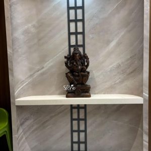 A stately 2-foot wooden Ganesha statue is elegantly positioned on a pristine white shelf against a backdrop of a sophisticated grey marbled wall. A striking black metal grid runs vertically behind the statue adding a contemporary design element to the traditional piece. Lord Ganesha is depicted in a seated posture radiating an aura of wisdom benevolence and auspiciousness. The rich dark tone and visible grain of the wood underscore the natural beauty and traditional craftsmanship of the statue making it a prominent feature in the room. Ganesha is presented with his characteristic elephant head large ears and four arms each holding symbolic objects. His upper hands grasp a conch shell and a disc representing divine power and protection. One of his lower hands holds his broken tusk while the other holds a modak signifying wisdom and the rewards of spiritual pursuit. The details of his ornamentation from his crown to his dhoti are meticulously carved. The statue rests on a beautifully sculpted lotus pedestal atop a rectangular base which features a prominent wheel motif adding layers of intricate detail and traditional symbolism. This image is ideally suited for an article discussing wooden Ganesha statues particularly in culturally rich cities like Ahmedabad Gujarat. The 2-foot size and robust wooden craftsmanship make it a significant and impactful piece for any home or office. Its elegant blend of traditional religious artistry with a modern display setting showcases its versatility and timeless appeal as a powerful symbol of good fortune success and spiritual harmony resonating deeply with local artistic traditions and contemporary interior design trends.