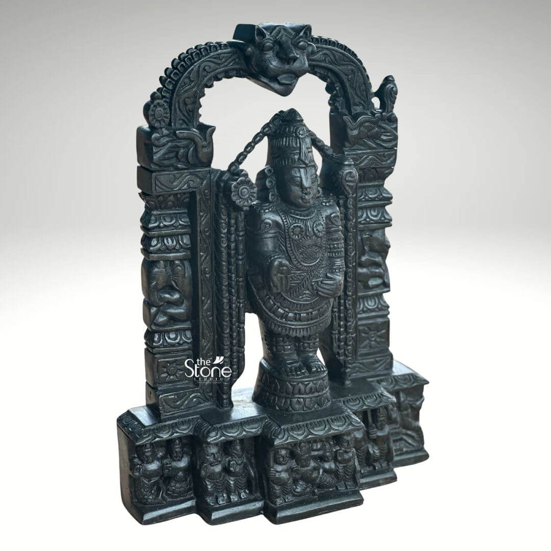 Tirupati Balaji Statue in Black Stone 18" 2 An exquisite 18-inch Venkateswara idol meticulously carved from black stone is presented in a slightly oblique front-right view against a plain light background making it an ideal centerpiece for a home temple. This stunning Balaji black stone statue captures Lord Venkateswara in his iconic standing pose emanating an aura of divine grace and profound power. The deep polished surface of the black stone provides a dramatic contrast enhancing the intricate details of the sculpture and its ornate architectural frame. Lord Venkateswara is depicted with his characteristic features including the prominent Urdhva Pundra on his forehead a majestic crown and various traditional ornaments such as multiple necklaces armlets and a sacred thread all finely detailed. His right hand is raised in the Abhaya Mudra offering blessings and reassurance to devotees while his left hand rests on his thigh symbolizing protection and prosperity. He stands on a lotus pedestal which is integrated into a multi-tiered base adorned with smaller figures and decorative carvings including devotees and mythological scenes all contributing to the grandeur of the piece. The intricate archway above him is equally detailed featuring divine figures floral motifs and a Kirtimukha at the apex. This18-inch black stone Venkateswara idol is a profound representation of Lord Venkateswara the preserver of the universe and is a perfect addition to a home temple. The exceptional craftsmanship in black stone not only makes it a visually striking piece but also embodies deep cultural and religious significance offering a powerful focal point for devotion and prayer. The rich detailing of the deity and his surrounding archway reflects centuries of tradition making it a truly remarkable and revered artifact that brings blessings and spiritual presence into any home.