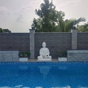 A serene eye-level photograph captures a luxurious outdoor swimming pool area featuring a magnificent 4-foot tall Fibre Buddha statue as the focal point. The statue rendered in a smooth matte white finish that mimics carved stone is seated in the traditional padmasana with hands in the dhyana mudra embodying profound tranquility and peace. It is positioned centrally on a raised stone platform directly at the water's edge appearing to watch over the crystal-clear blue tiled pool. The calm expression and detailed robes of the statue showcase the high craftsmanship of its lightweight fibre construction making it a substantial and durable piece of garden art. The background is dominated by a substantial boundary wall constructed from dark brown textured brick cladding offering a rich contrast to the white statue. This wall is punctuated by two light-gray granite pillars topped with modern globe-shaped outdoor lamps. To the left and right of the Buddha statue integrated into the same raised stone plinth are two rectangular white planters containing small manicured green shrubs adding a touch of nature and symmetry to the shrine-like setting. Above the wall lush green trees and a palm frond rise against a cloudy sky creating a private tropical oasis feel. The placement of this 4-foot Fibre Buddha statue transforms the swimming pool area into a tranquil retreat perfect for relaxation and meditation. The statue's size is impressive but not overwhelming creating a harmonious balance with the pool and the surrounding landscaping. Its elevated position at the water's edge makes it a stunning visual element visible from all parts of the pool deck and its light color stands out beautifully especially during the day. This image serves as an ideal inspiration for anyone looking to incorporate elegant Eastern-inspired elements into their swimming pool decor.